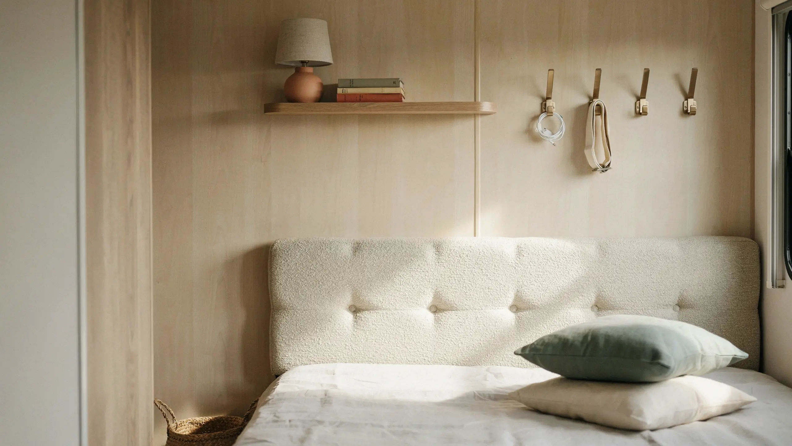 Minimalist floating shelf and wall hooks installed above a bed with a tufted boucle headboard, holding a terracotta lamp, books, and hanging accessories in a small bedroom.