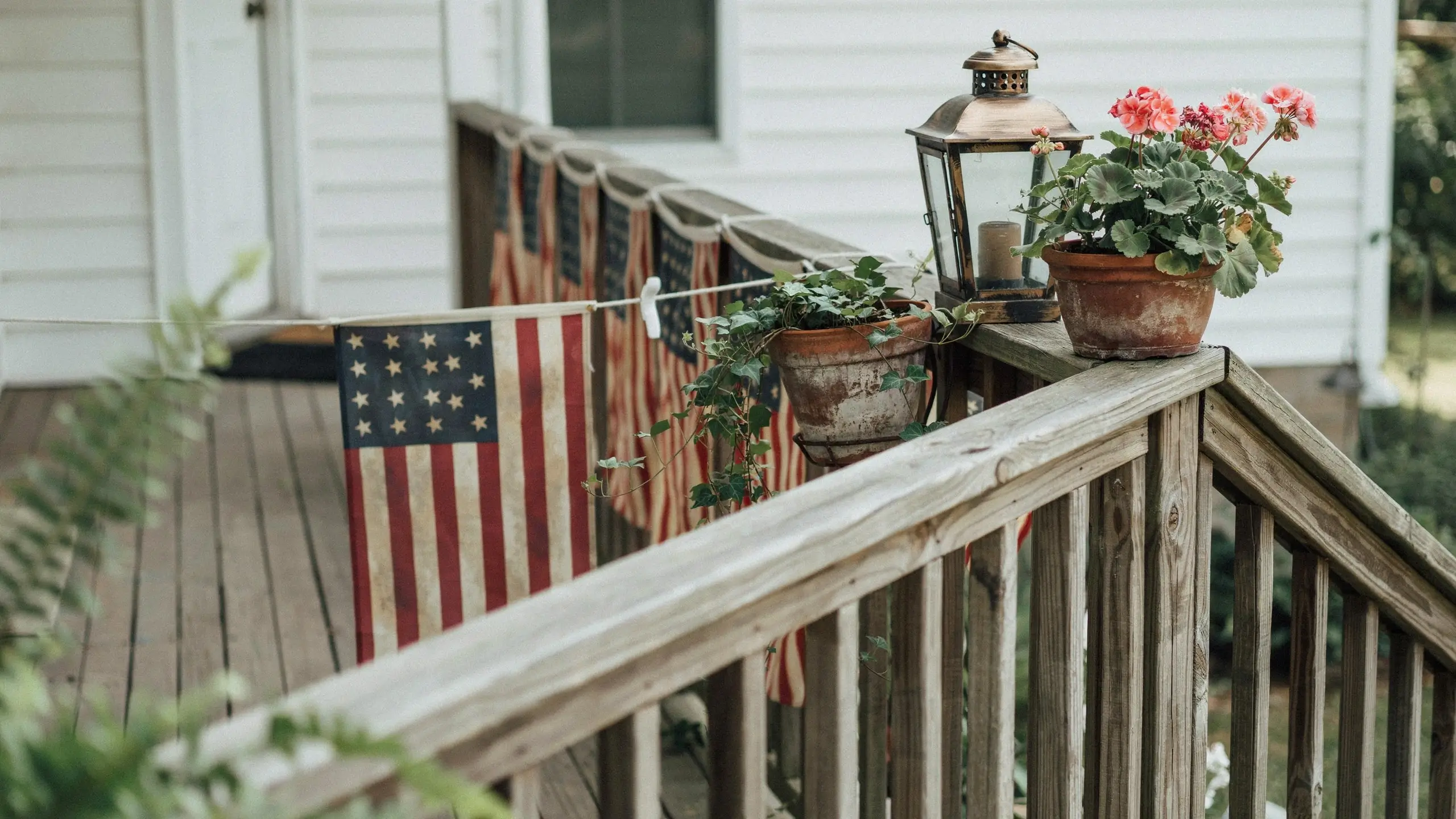 Vintage-style American flag banners lining a wooden porch railing with flowers and a rustic lantern