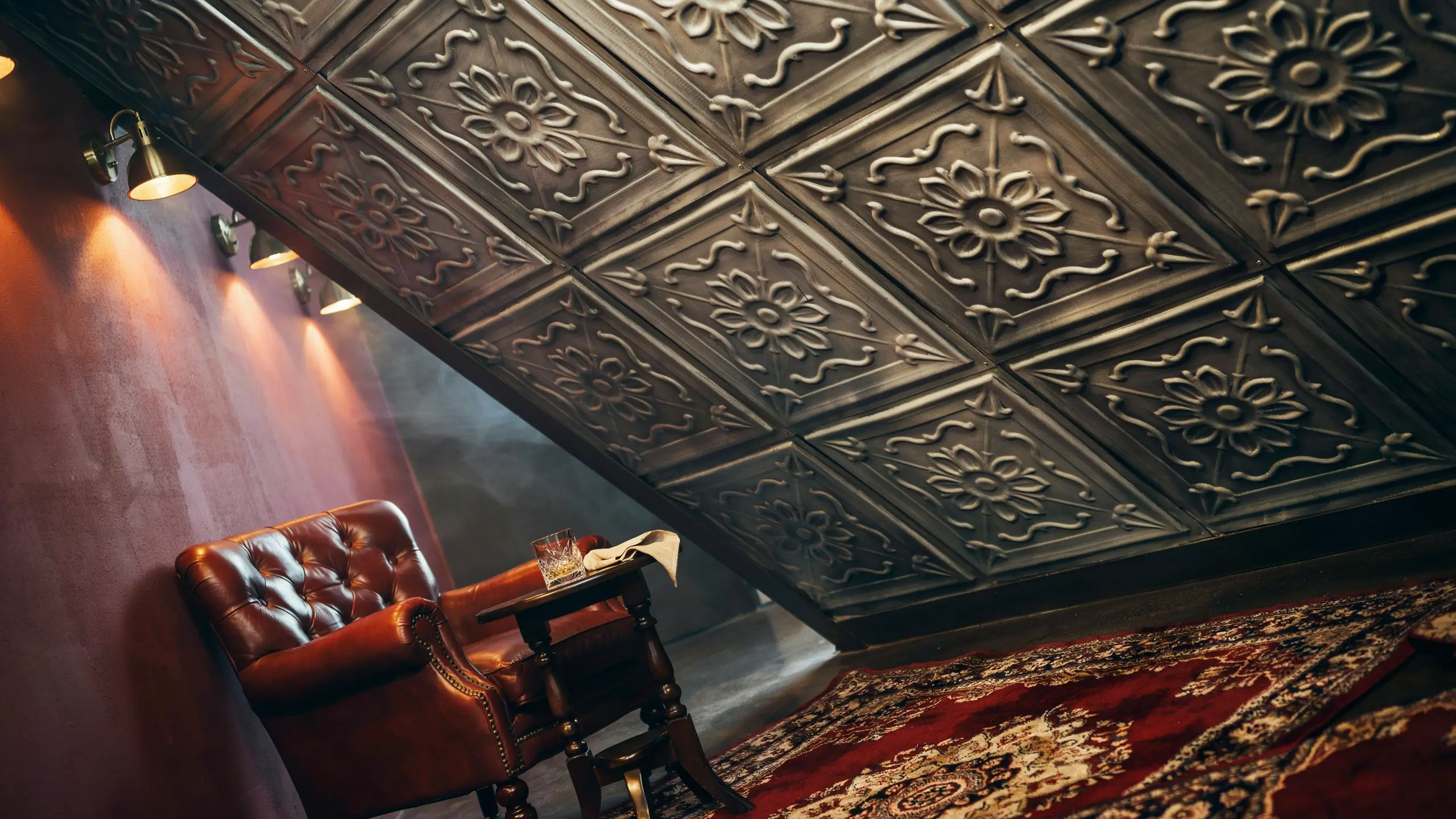Dark antique-style embossed tin ceiling panels on a sloped ceiling above a brown tufted leather armchair with warm wall sconce lighting and a patterned rug