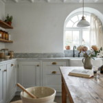17 Vintage Farmhouse Kitchen Ideas With a Modern Twist You’ll Want to Pin Vintage farmhouse kitchen with white cabinets, marble counters, open wood shelves, arched window, and cat on rustic table.