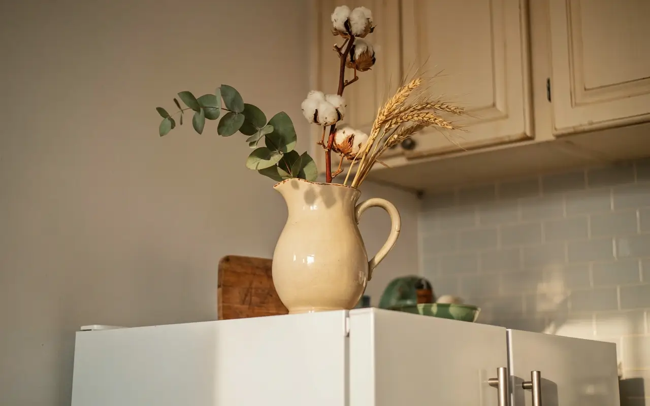 Vintage cream pitcher with eucalyptus, cotton stems, and dried wheat displayed above a white refrigerator.