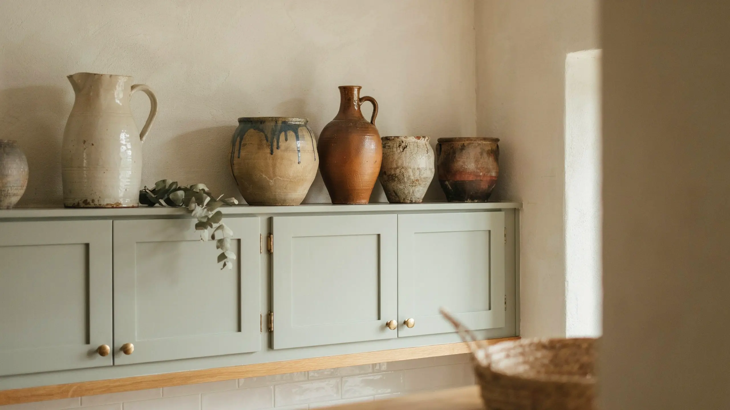 Vintage pottery and ceramic crocks displayed above sage green kitchen cabinets with trailing greenery.