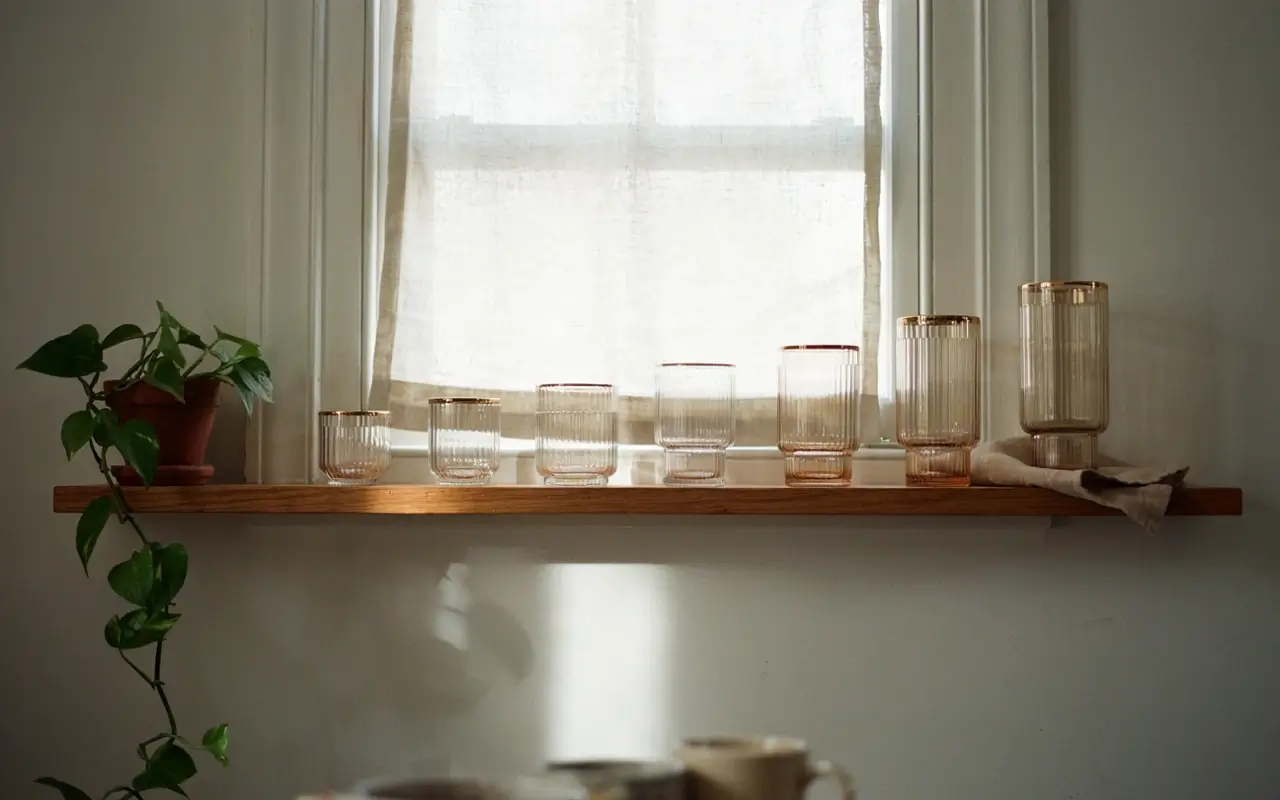 Vintage ribbed glassware stacked by height on a floating wood shelf in front of a softly lit kitchen window.