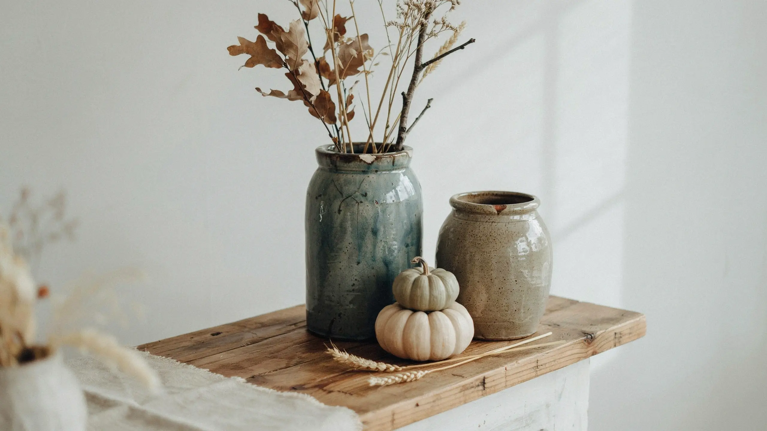 Two rustic stoneware crocks featuring a tall blue-grey glazed vase filled with dried oak leaves and wheat stalks, and a shorter speckled beige jar, sitting on a wooden shelf with stacked white and green mini pumpkins.