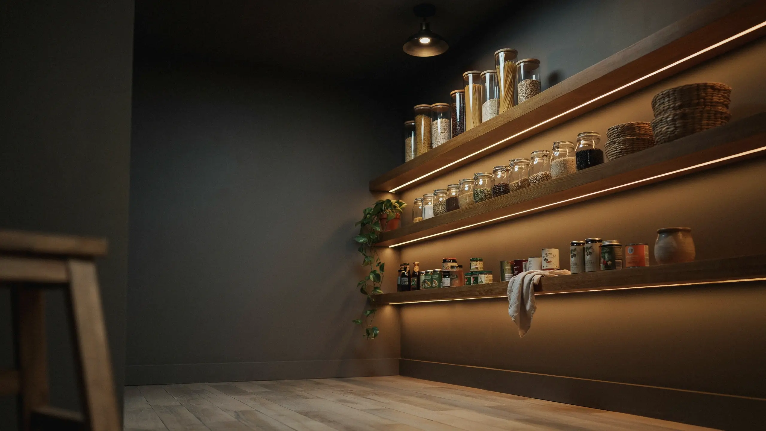 Dark modern walk-in pantry featuring an upgraded LED strip lighting system under wooden floating shelves, illuminating glass jars filled with dry pantry staples like pasta, grains, and canned goods.