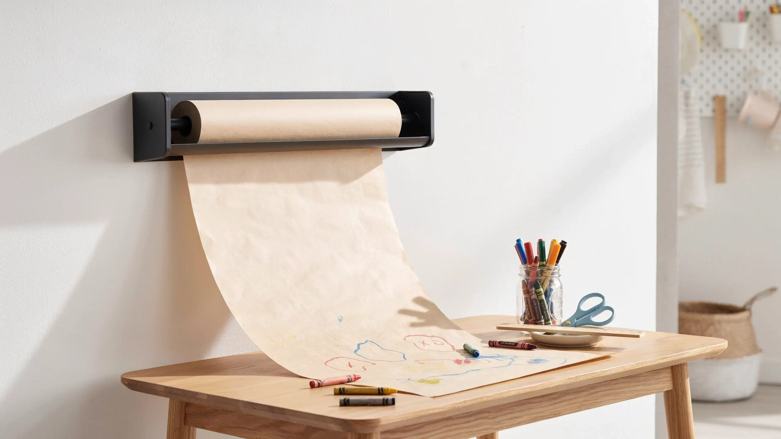 "Wall-mounted kraft paper roll dispenser with drawing paper draped over a light wood table, surrounded by colorful crayons, markers in a glass jar, and scissors for kids' art projects." 
