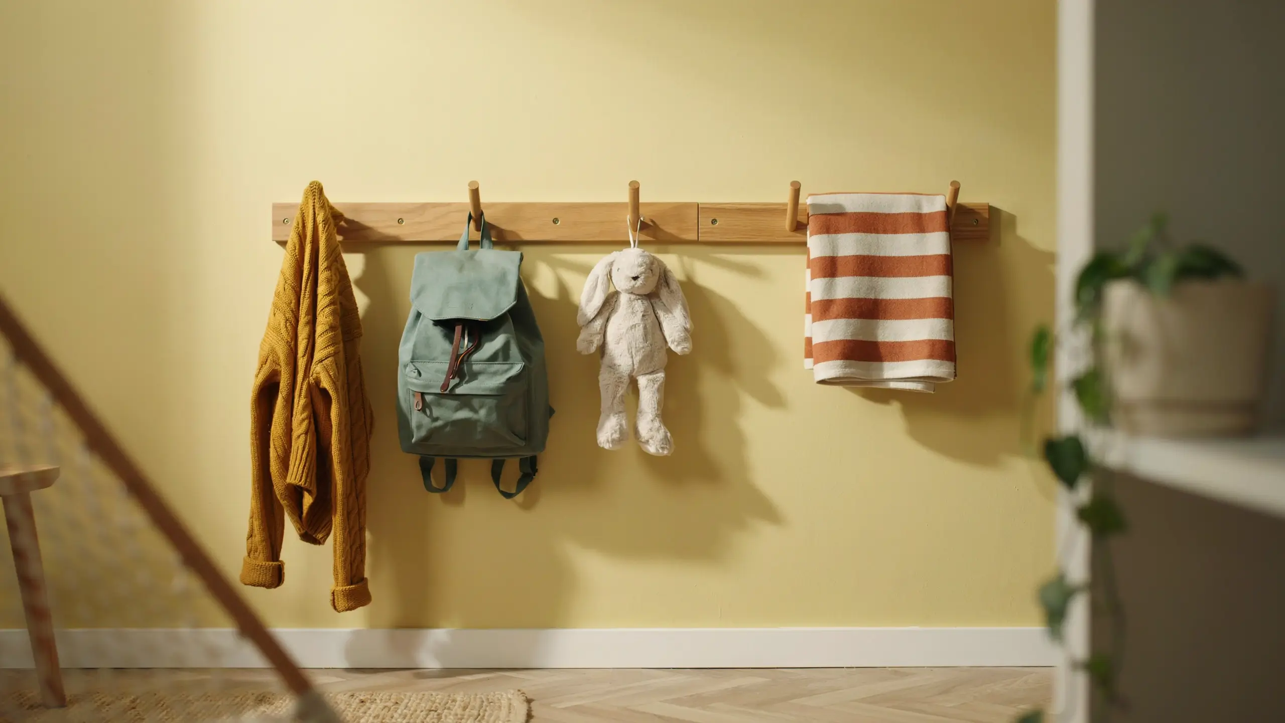 A warm-toned wall-mounted wooden peg rail installed in a yellow hallway, holding a mustard-yellow cable-knit sweater, a sage-green canvas backpack, a fluffy white plush bunny toy, and a rust-striped towel as an efficient, space-saving storage solution for entryway clutter. 