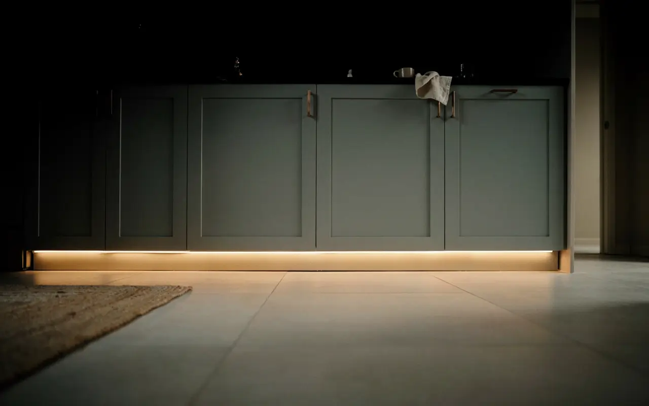 Warm LED strip under sage kitchen cabinet toe-kick creates a soft floating effect on the tiled floor.