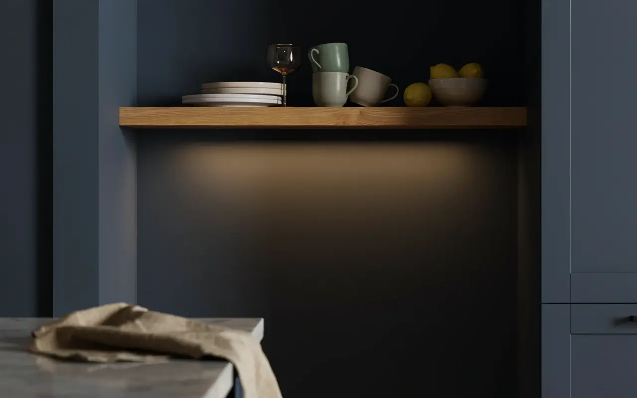 Hidden warm LED strip under a floating wood shelf in a dark blue kitchen with mugs, plates, and lemons.