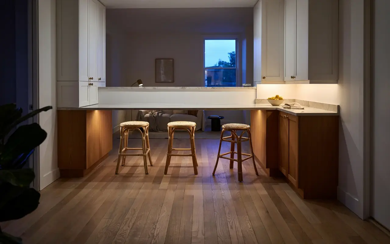 Warm LED strip glowing beneath a kitchen peninsula overhang with rattan stools and white cabinets.