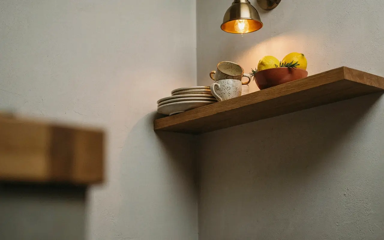 Warm pendant light above a wooden open kitchen shelf with stacked plates, mugs, and lemons in a bowl.