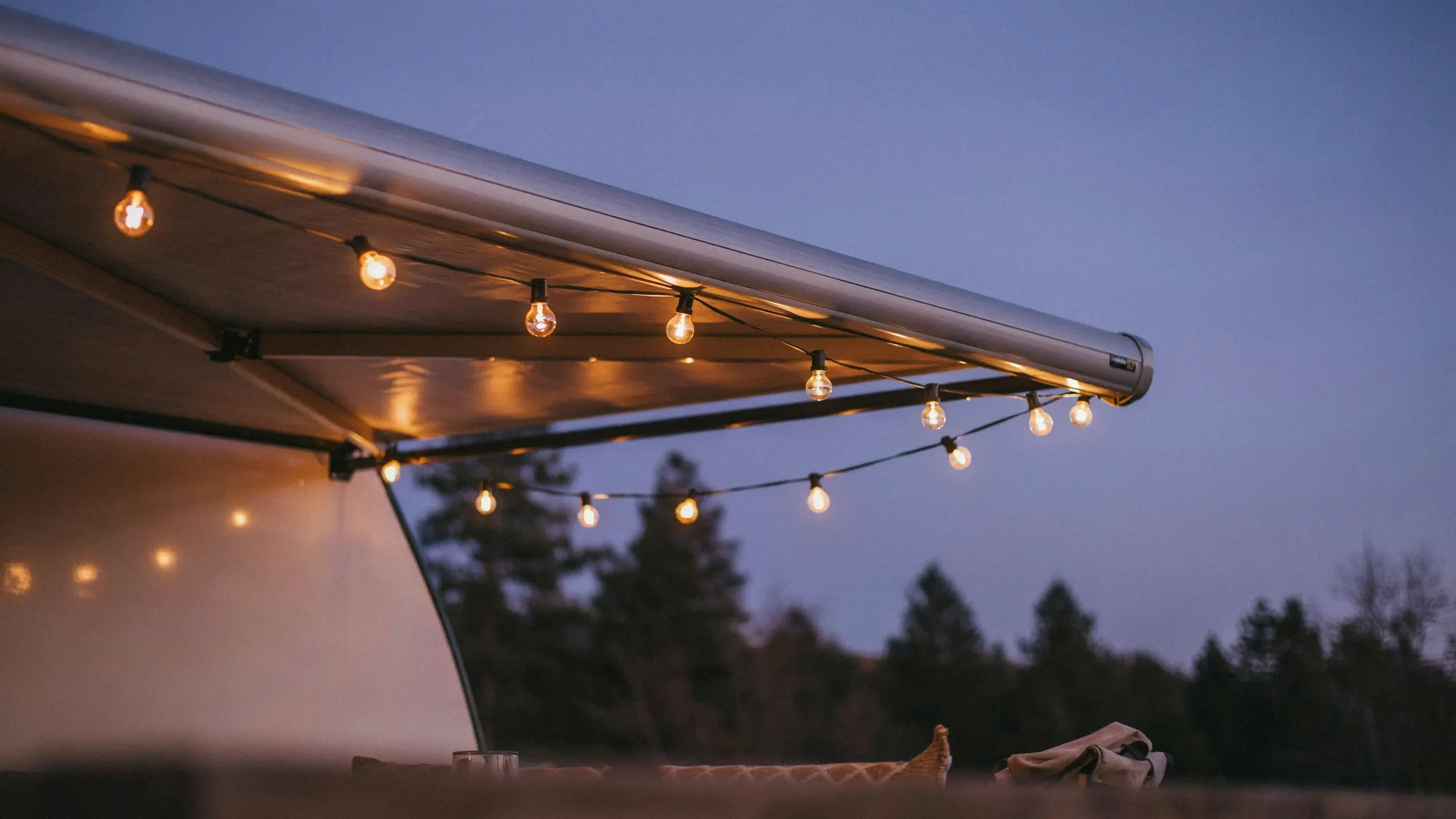 Warm white globe string lights hanging along a camper awning at dusk, creating a cozy outdoor camping ambiance. 
