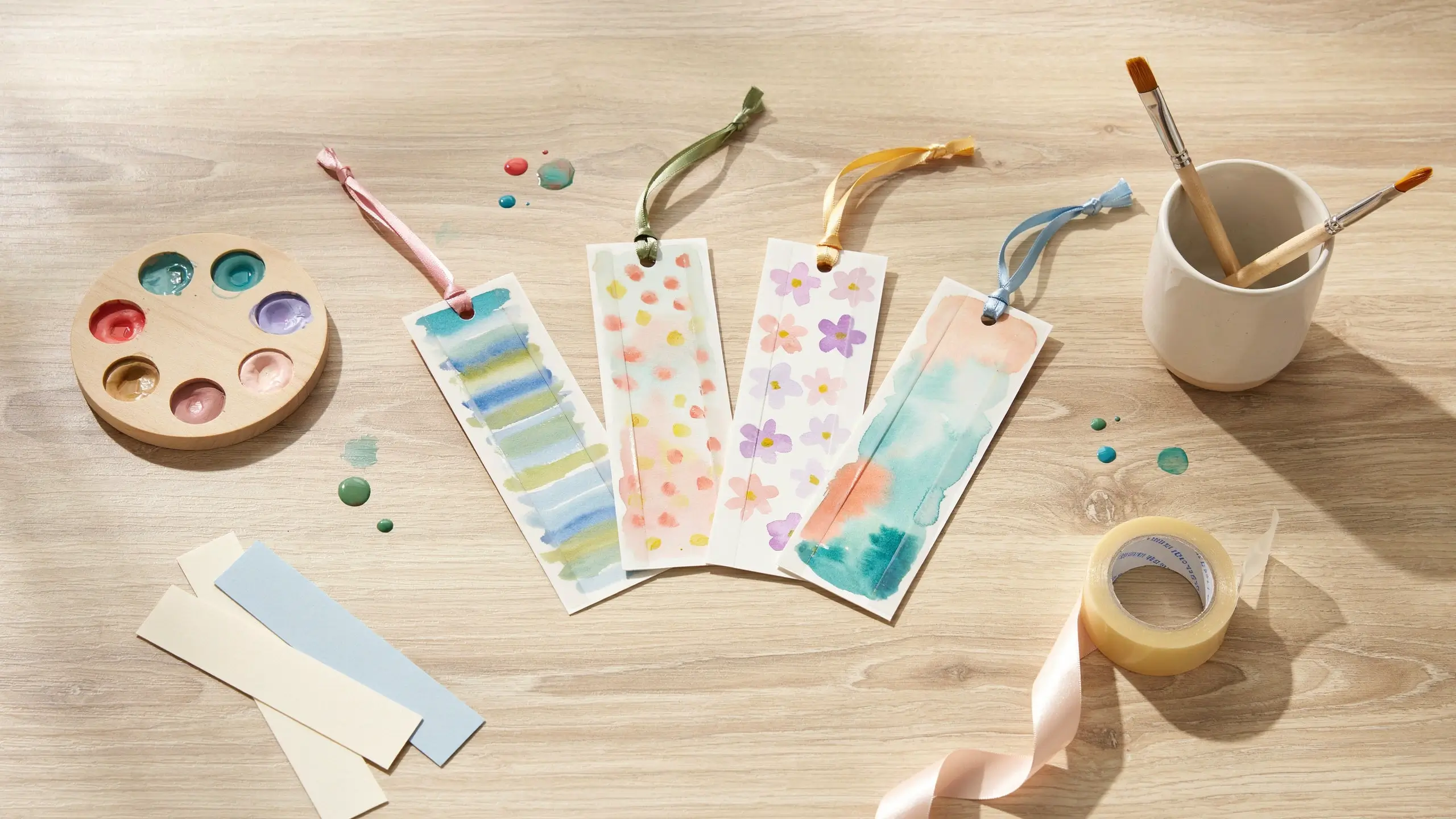 Handmade watercolor bookmarks with ribbons, paintbrushes, and a paint palette on a wooden table.