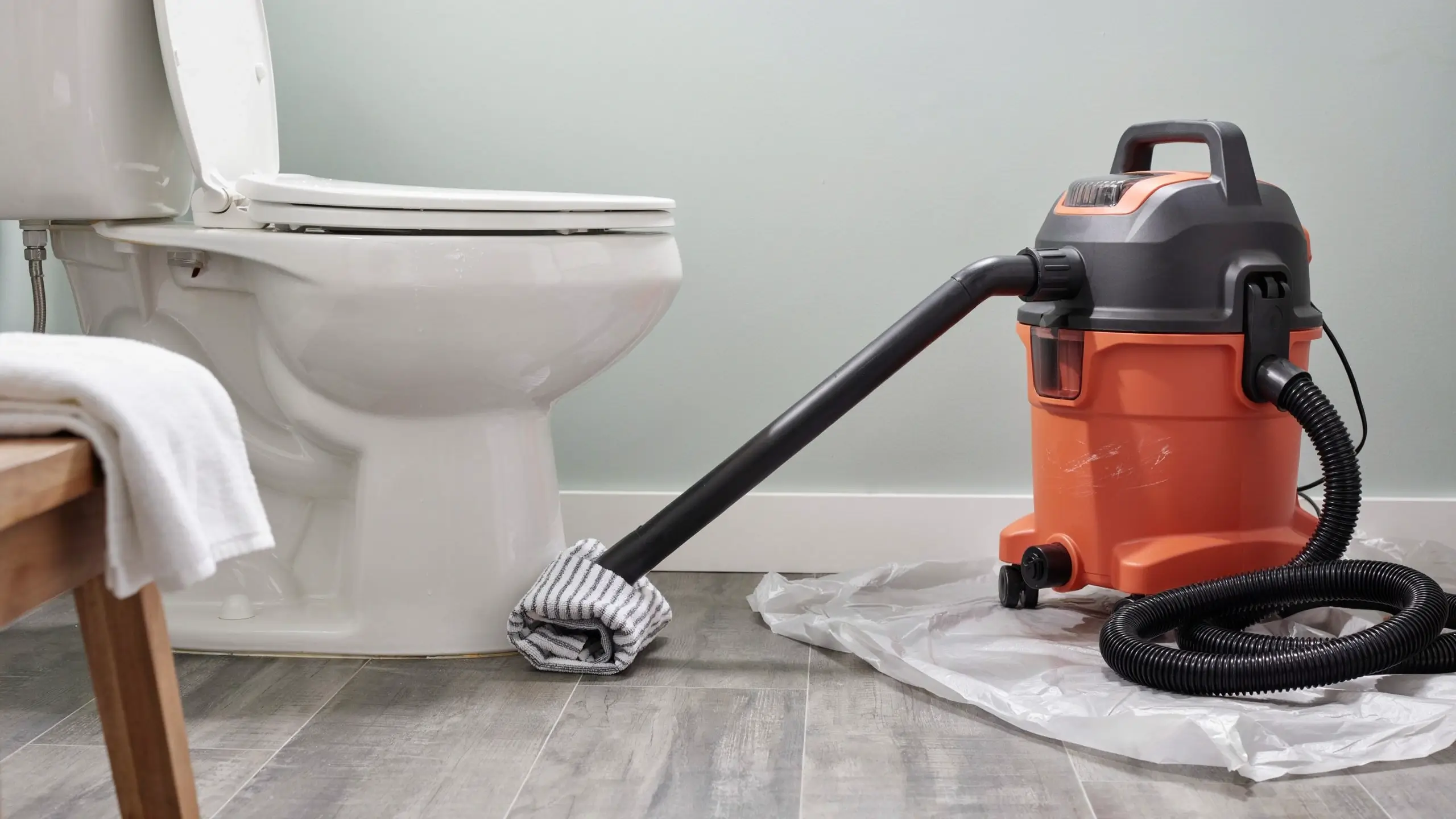 Orange and gray wet/dry shop vacuum cleaner with a black corrugated hose, resting on a white drop cloth, using a white cloth attachment to clean the base of a white porcelain toilet in a bathroom.