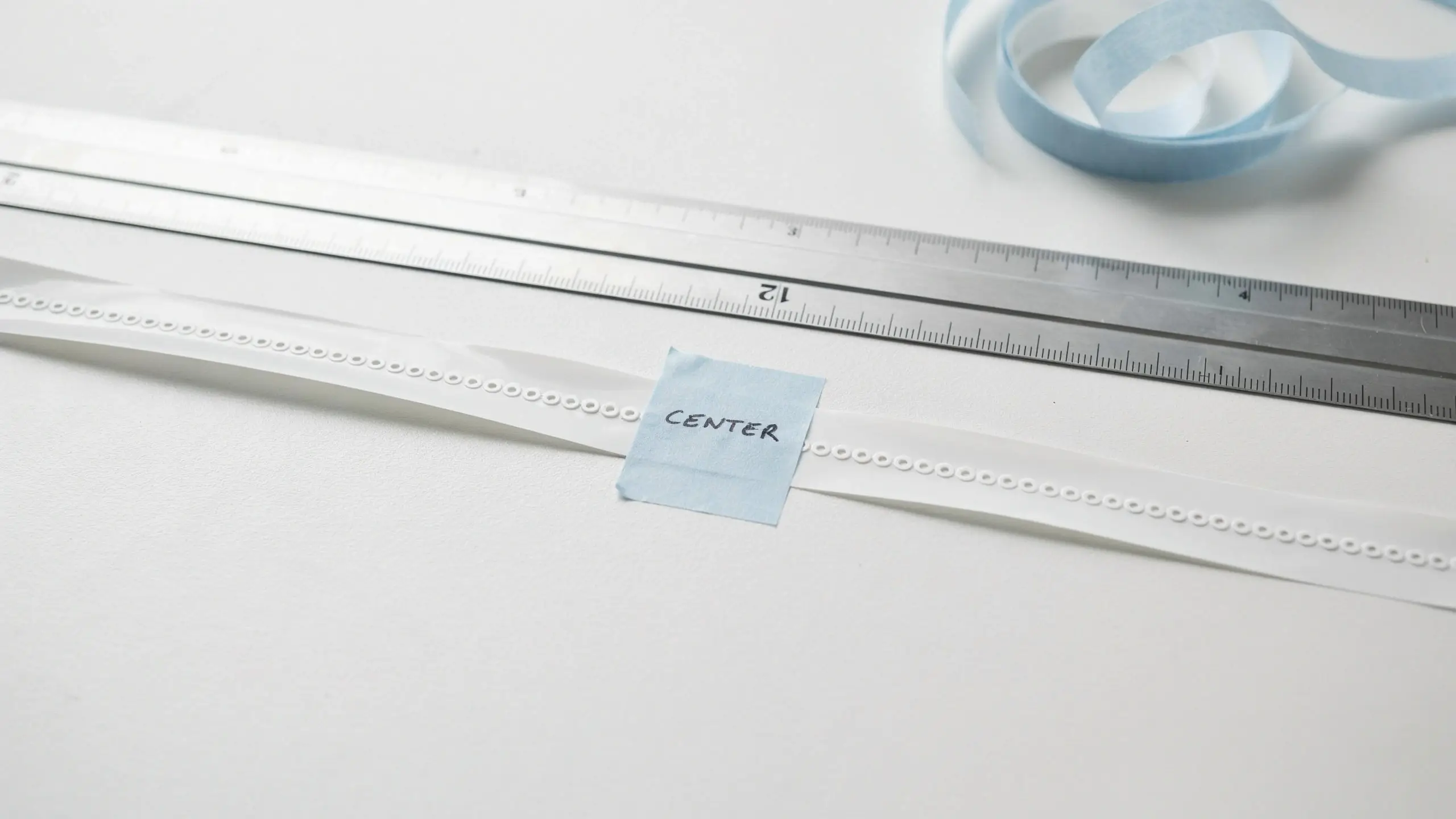 A white perforated craft decorating strip marked precisely in the middle with a piece of blue tape labeled "CENTER", placed next to a metal ruler to ensure accurate measurement for symmetrical decoration.
