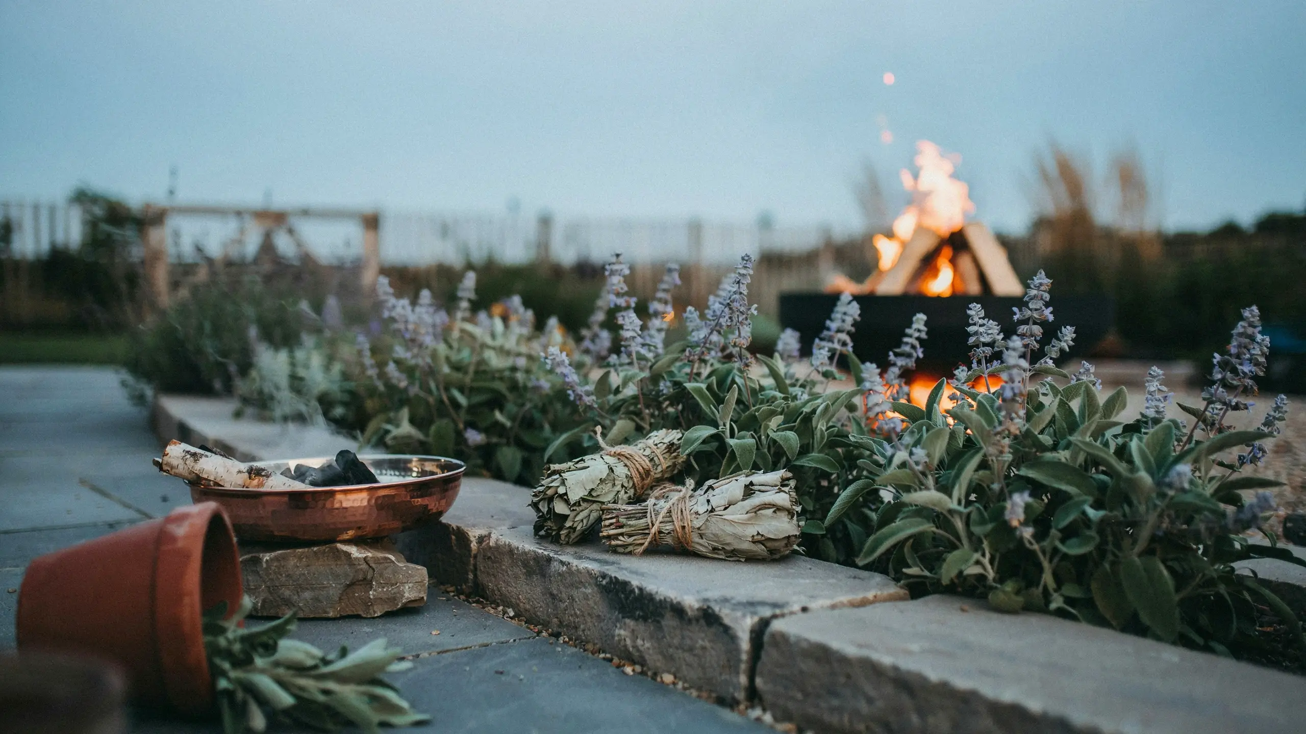 Dried white sage smudge sticks bound with twine resting on a stone patio ledge, next to a copper burning bowl and an outdoor fire pit during twilight. 