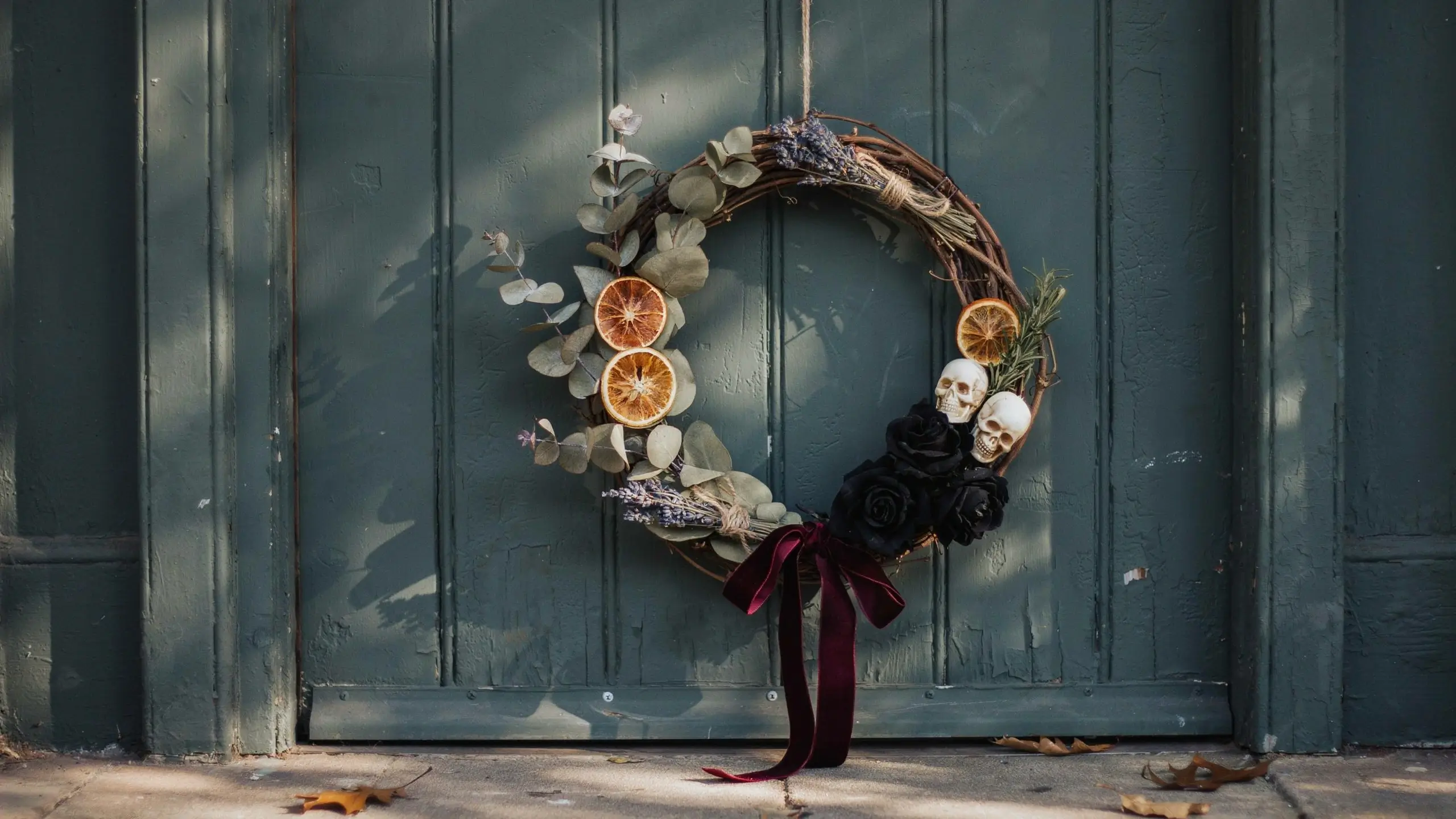 Handcrafted witchy dried botanical wreath on a dark green wooden door, featuring eucalyptus, dried orange slices, lavender, black roses, and small white skulls tied with a burgundy velvet bow. 