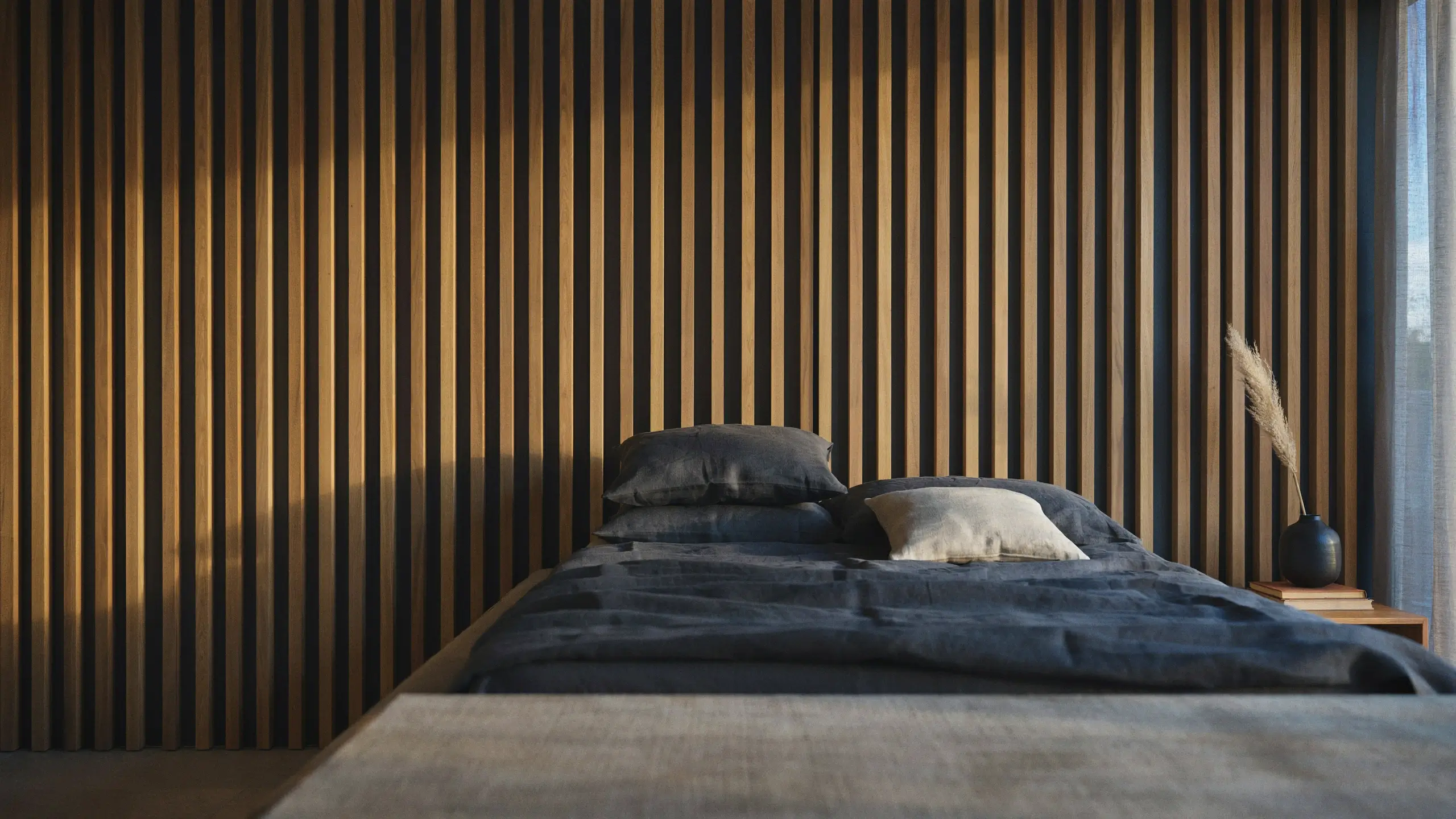 Modern bedroom interior featuring a vertical wood slat or fluted panel accent wall, dark charcoal bedding, and warm natural sunlight casting dramatic vertical shadows.
