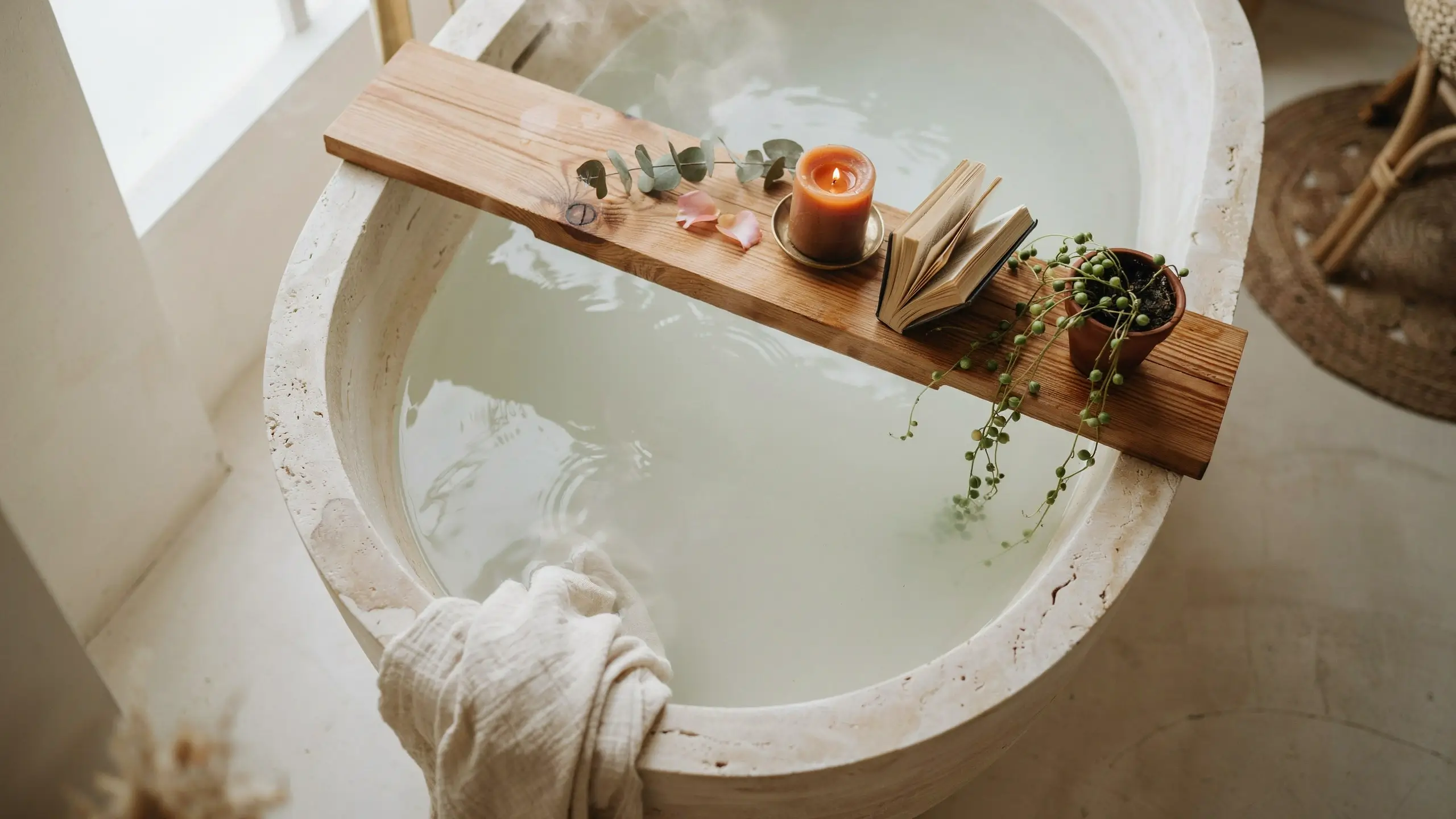 A rustic wooden bath caddy or tray resting across a white stone bathtub filled with steamy milky water, holding a lit orange candle, an open book, rose petals, eucalyptus, and a trailing string-of-pearls potted plant.