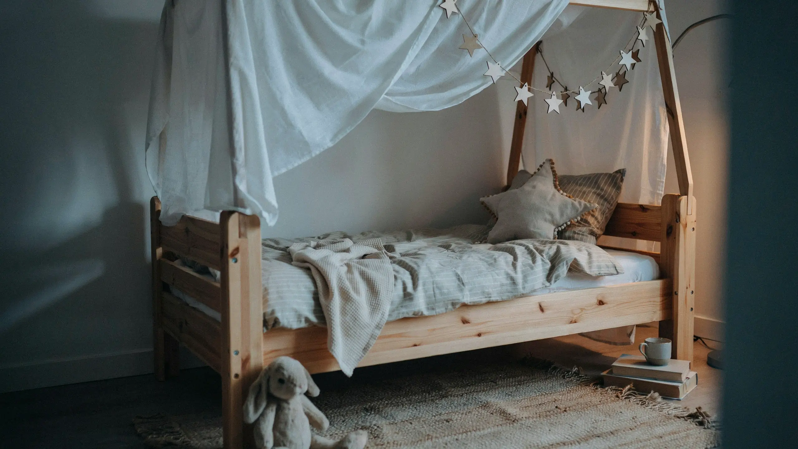 A cozy pine wood house-frame bed for a toddler, draped in a white fabric canopy and decorated with a star garland, featuring striped bedding and a plush bunny toy resting on the floor in a minimalist nursery. 