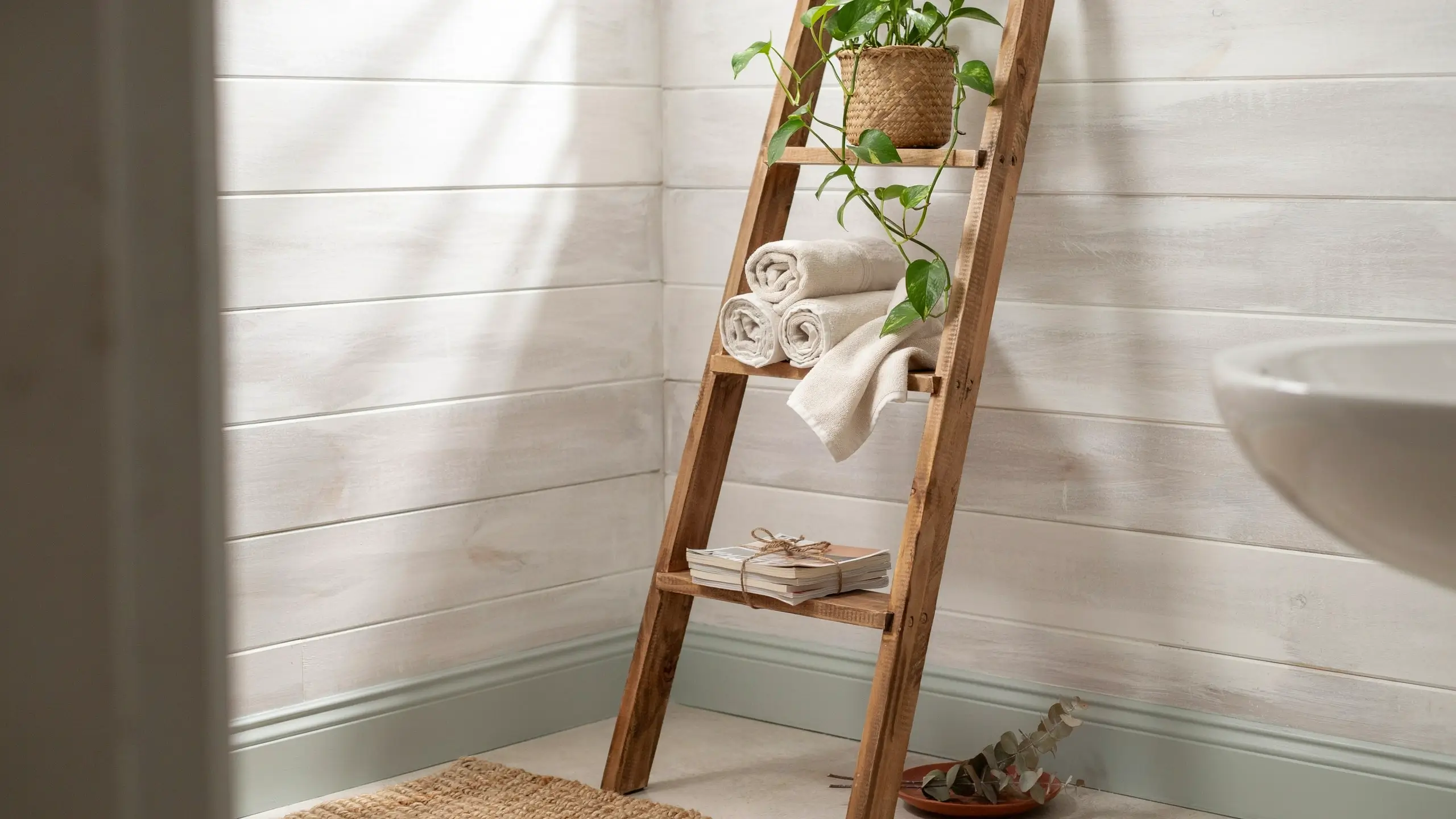 Freestanding rustic wooden ladder shelf holding rolled white towels, a potted plant, and books in a bright farmhouse bathroom