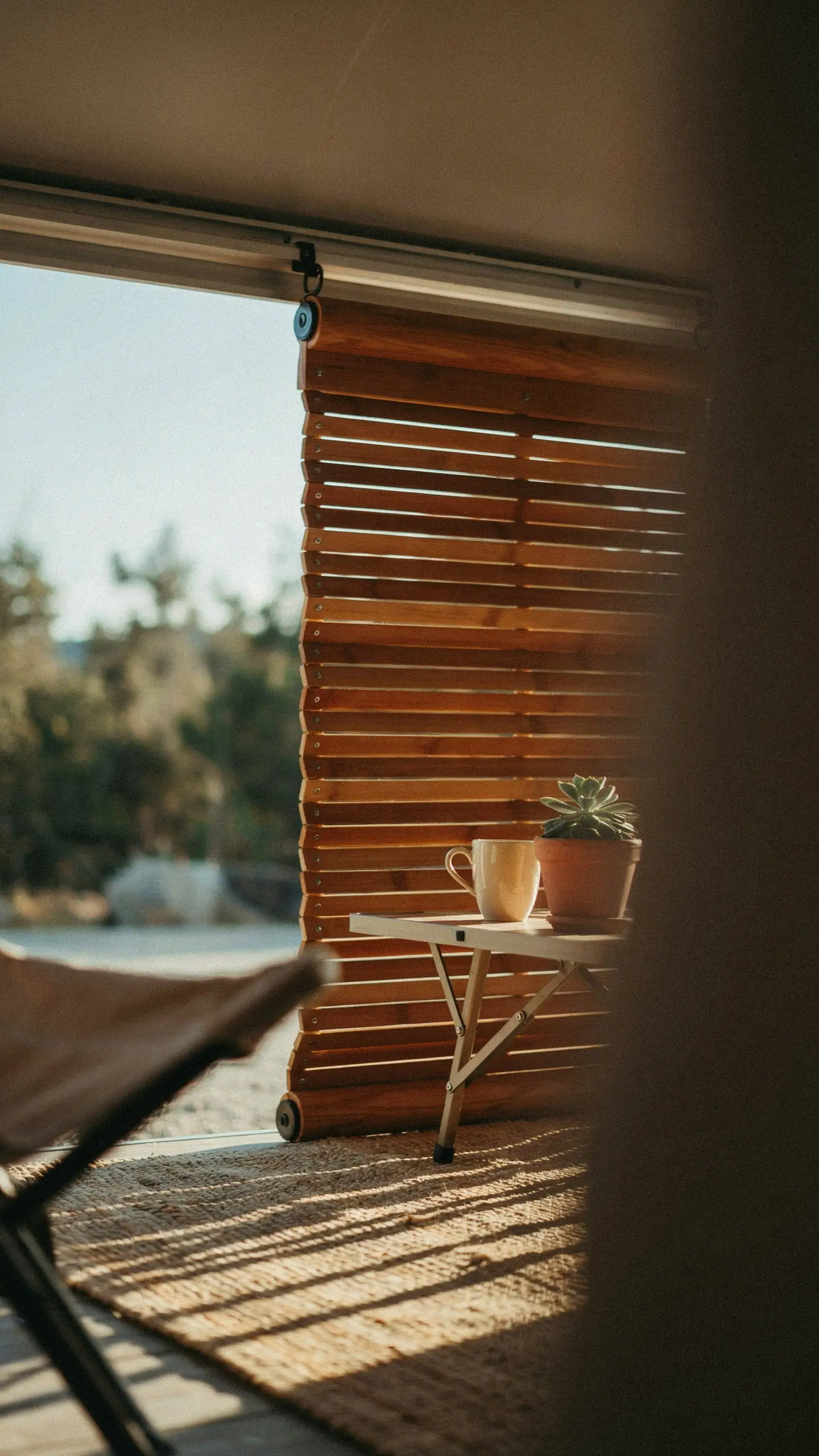 Wooden slatted privacy screen attached to awning arms filtering sunlight over a small outdoor table with a coffee mug and potted succulent. 