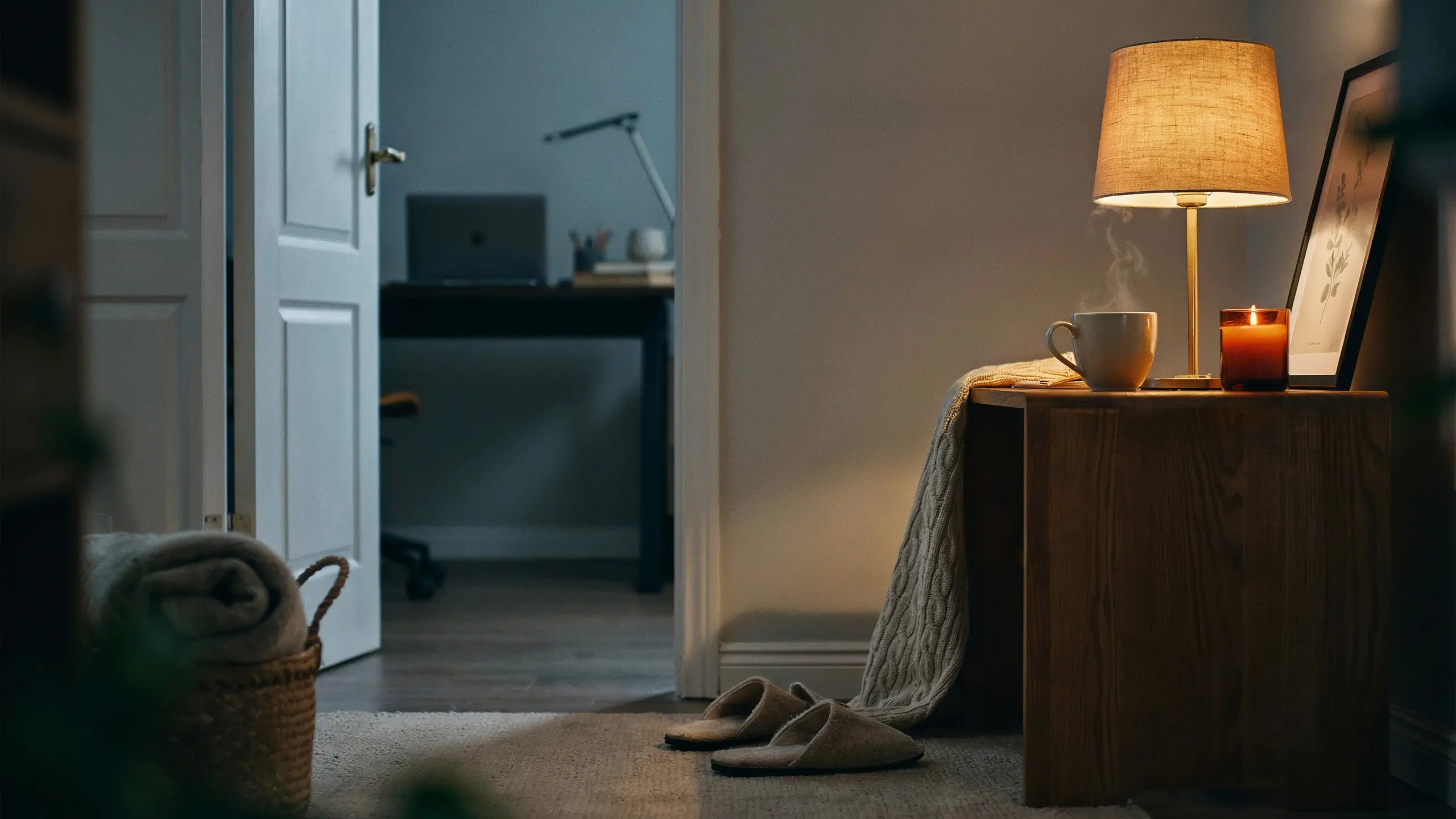 Warm lamp illuminating steaming coffee mug, candle, and slippers next to open home office door for evening wind-down routine.