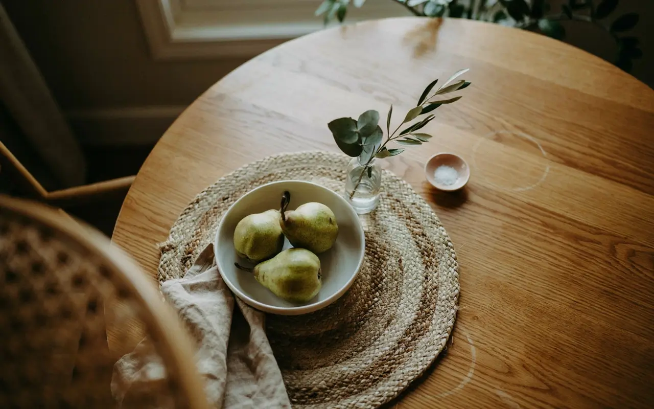 Woven placemat anchoring a bowl of pears and greenery on a small round wood dining table.
