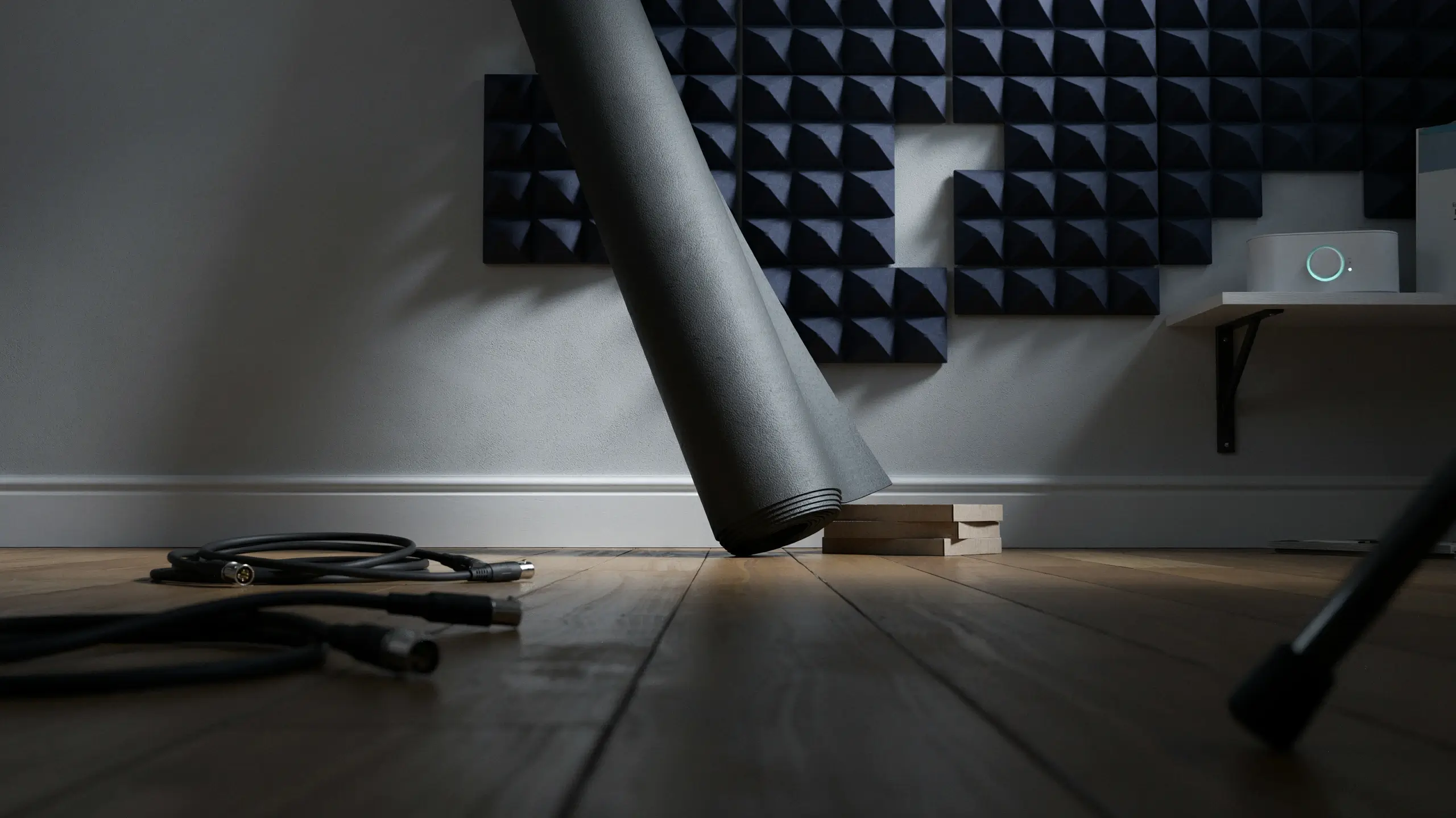 Rolled black acoustic soundproofing mat leaning against wall-mounted pyramid foam panels, with XLR cables and a speaker device on a wooden floor.