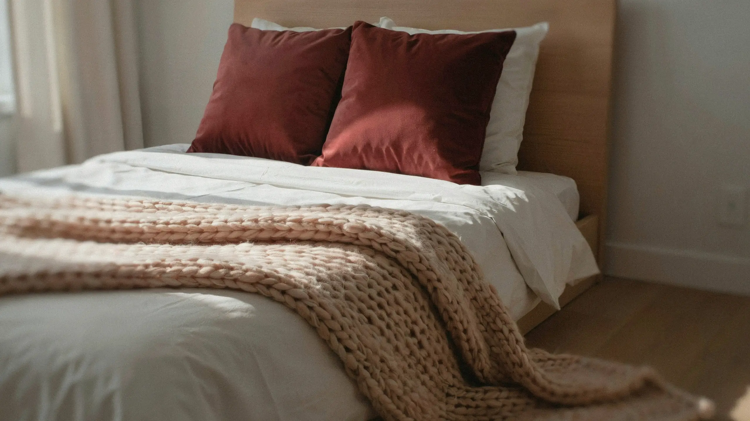 Rust-red velvet pillows layered against white bedding with a heavy chunky-knit beige throw blanket draped over the foot of the bed.