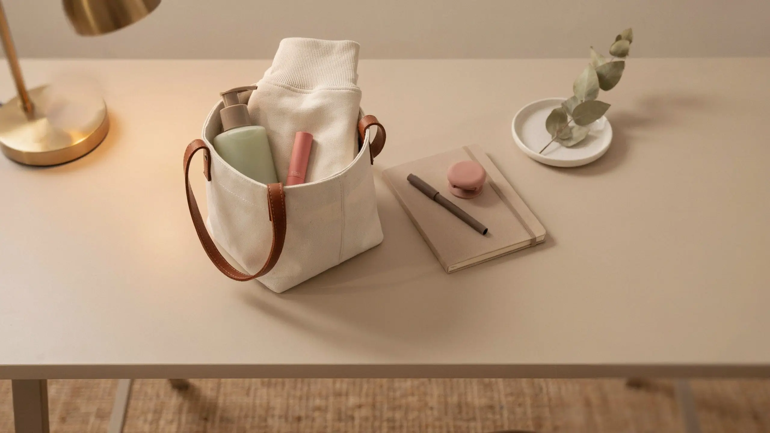 A modern, minimalist Easter basket idea for teens and tweens featuring a cream canvas tote bag holding a folded white sweatshirt, a green skincare bottle, and pink lip gloss, sitting on a desk next to a beige notebook and a small pink macaron.