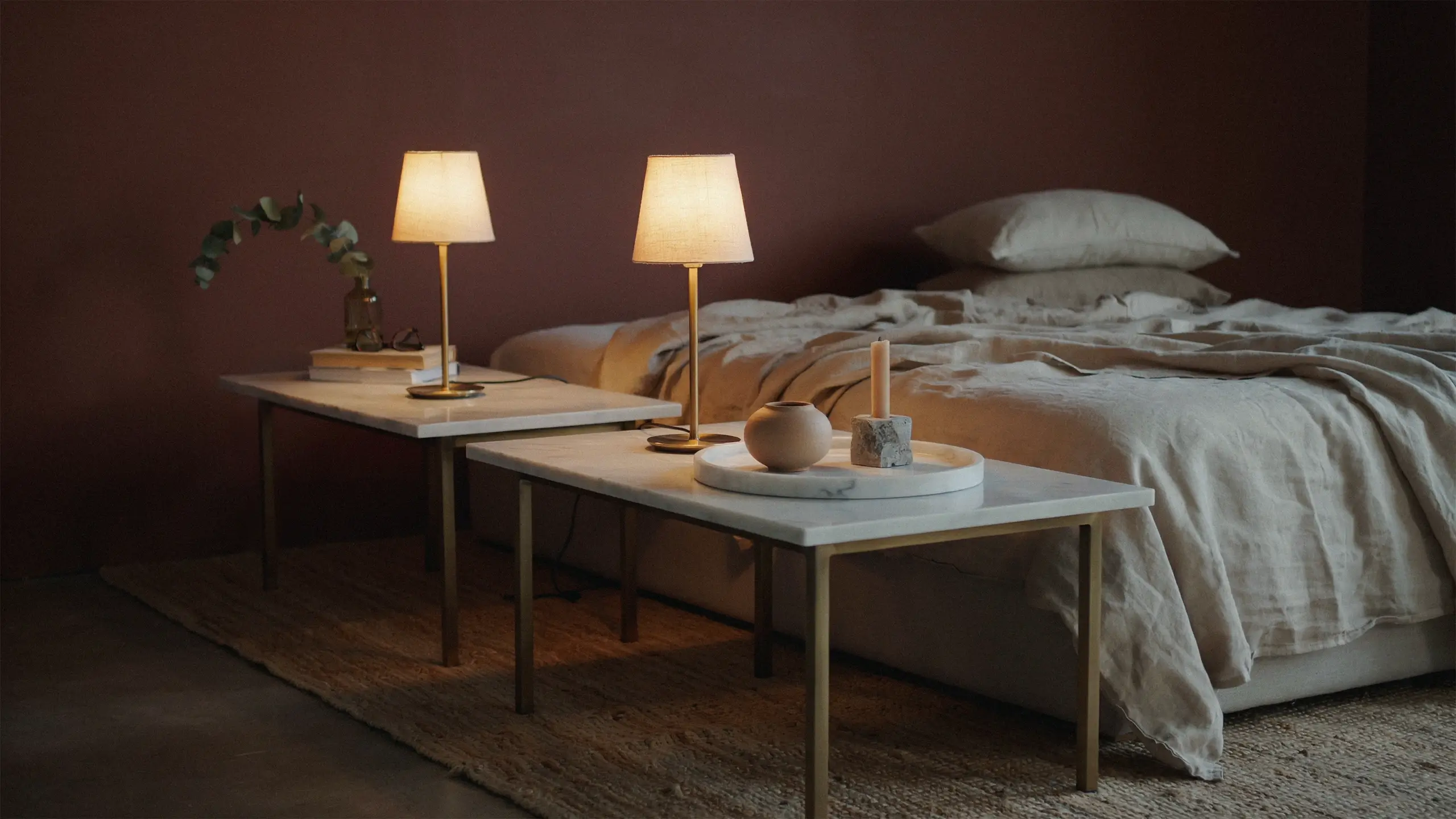 Warm, cozy bedroom featuring two budget-friendly white marble top side tables with gold legs, illuminated by warm lamps, alongside an unmade bed with soft linen bedding, a jute rug, and natural stone accents.