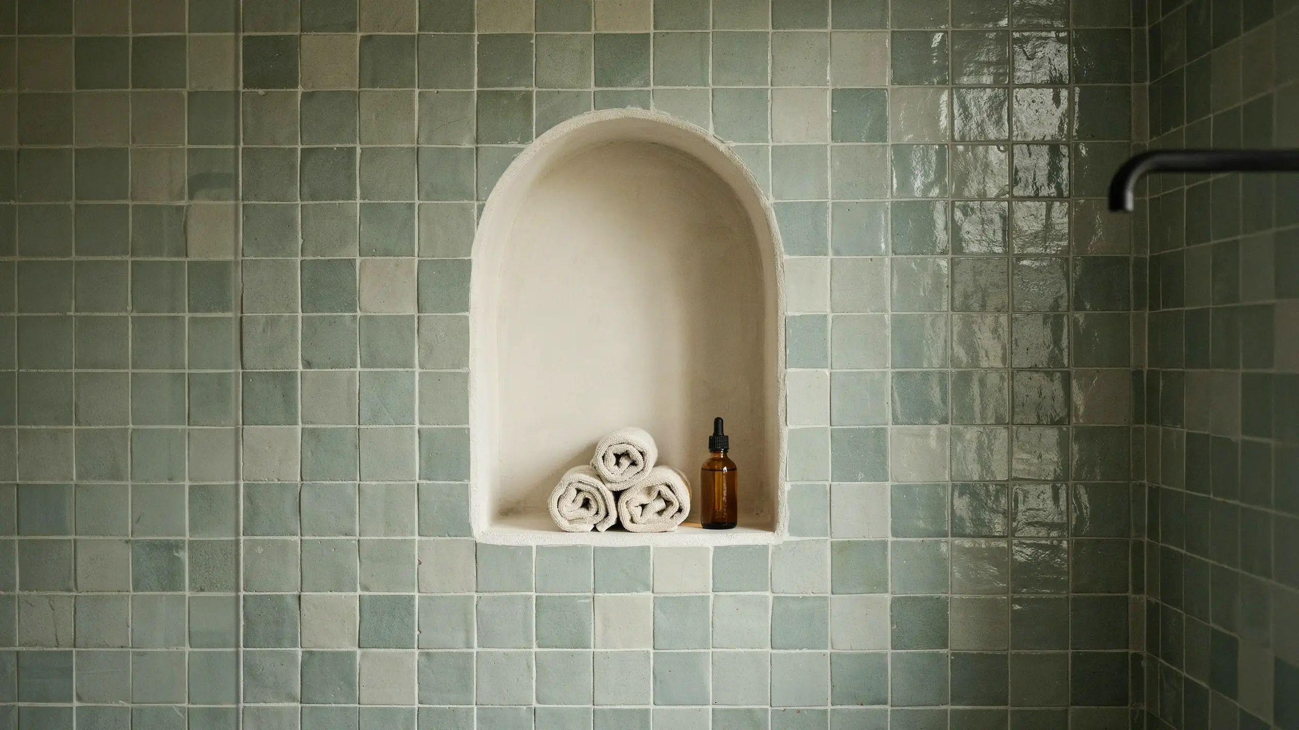 Arched shower niche framed with square sage green ceramic tiles, featuring rolled white towels and an amber glass essential oil bottle.