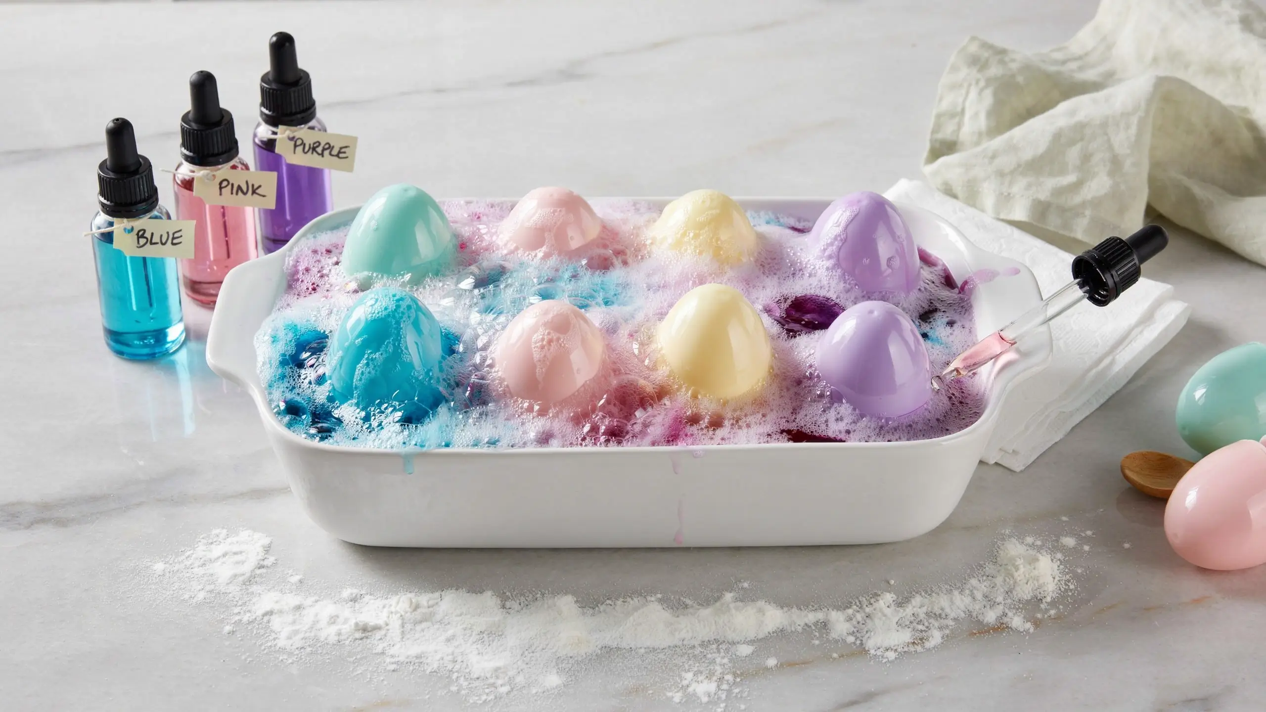 Pastel-colored Easter eggs fizzing in a white ceramic dish with a colorful baking soda and vinegar reaction using dropper bottles.