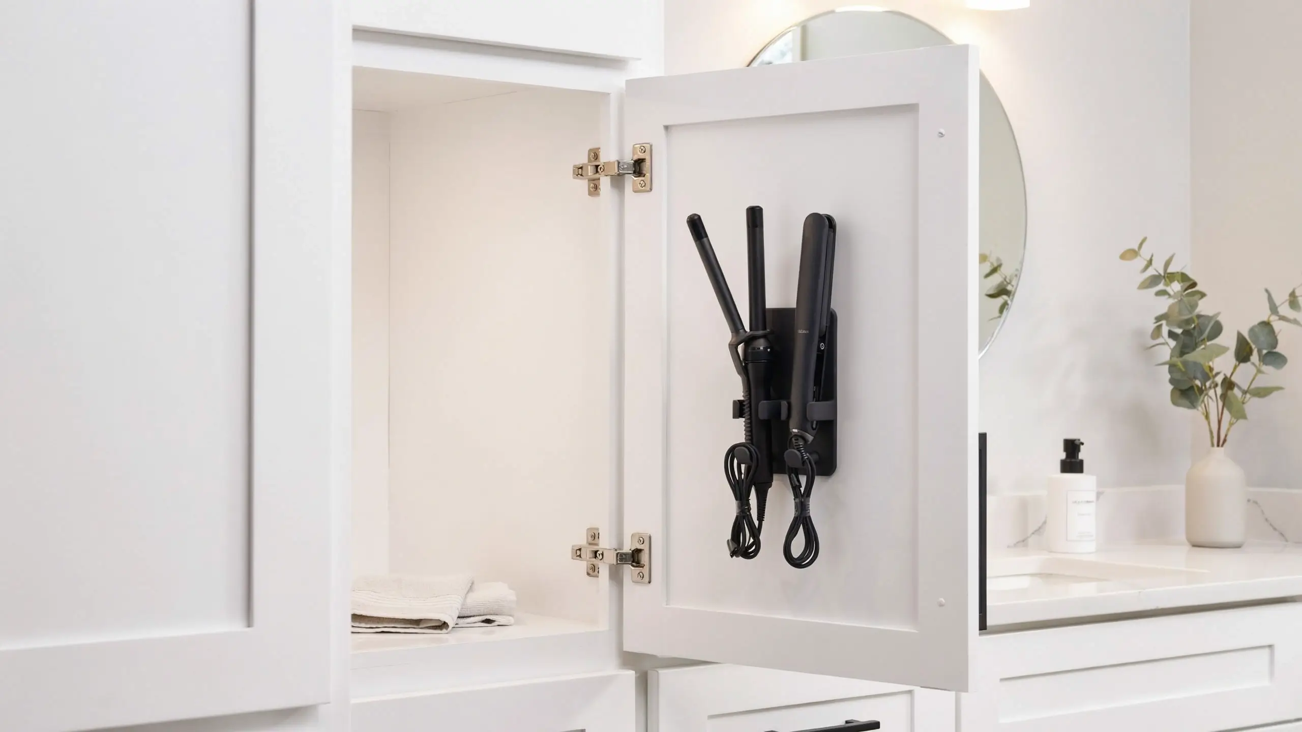 White bathroom vanity cabinet door featuring a black, space-saving hair tool organizer mounted on the inside, securely holding a flat iron and a curling wand to free up counter space.