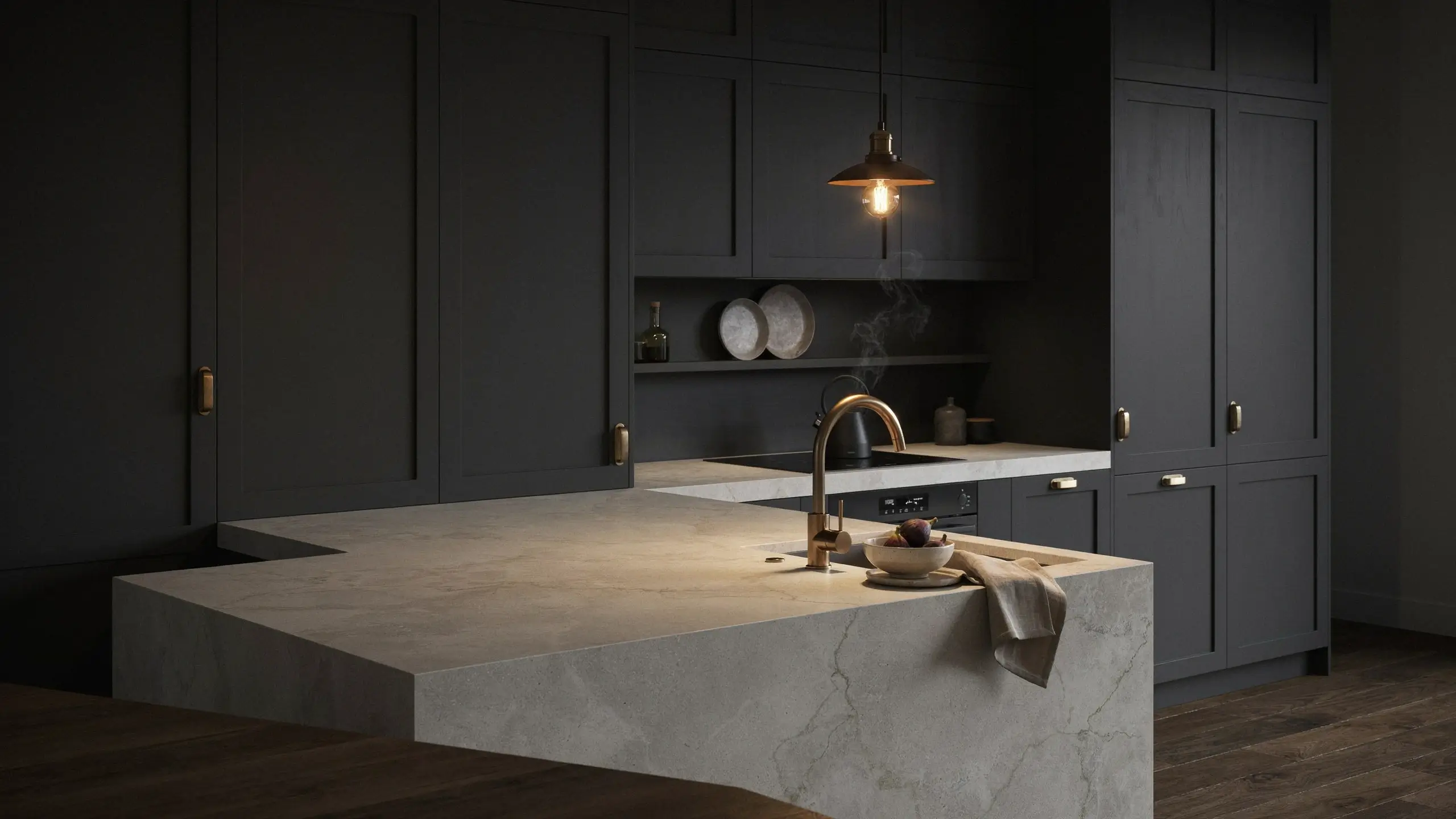 A moody, high-end kitchen featuring dark matte shaker-style custom cabinet fronts with brass hardware. The bespoke look is centered around a light-veined stone island equipped with a brushed brass faucet, a bowl of figs, and a teakettle emitting steam under warm pendant lighting.