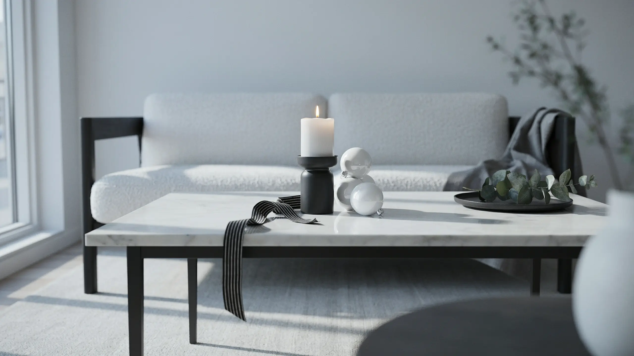 A minimal black and white Christmas palette featuring a glowing white pillar candle, silver spherical ornaments, and a black striped ribbon on a marble coffee table, placed in front of a modern white boucle sofa.