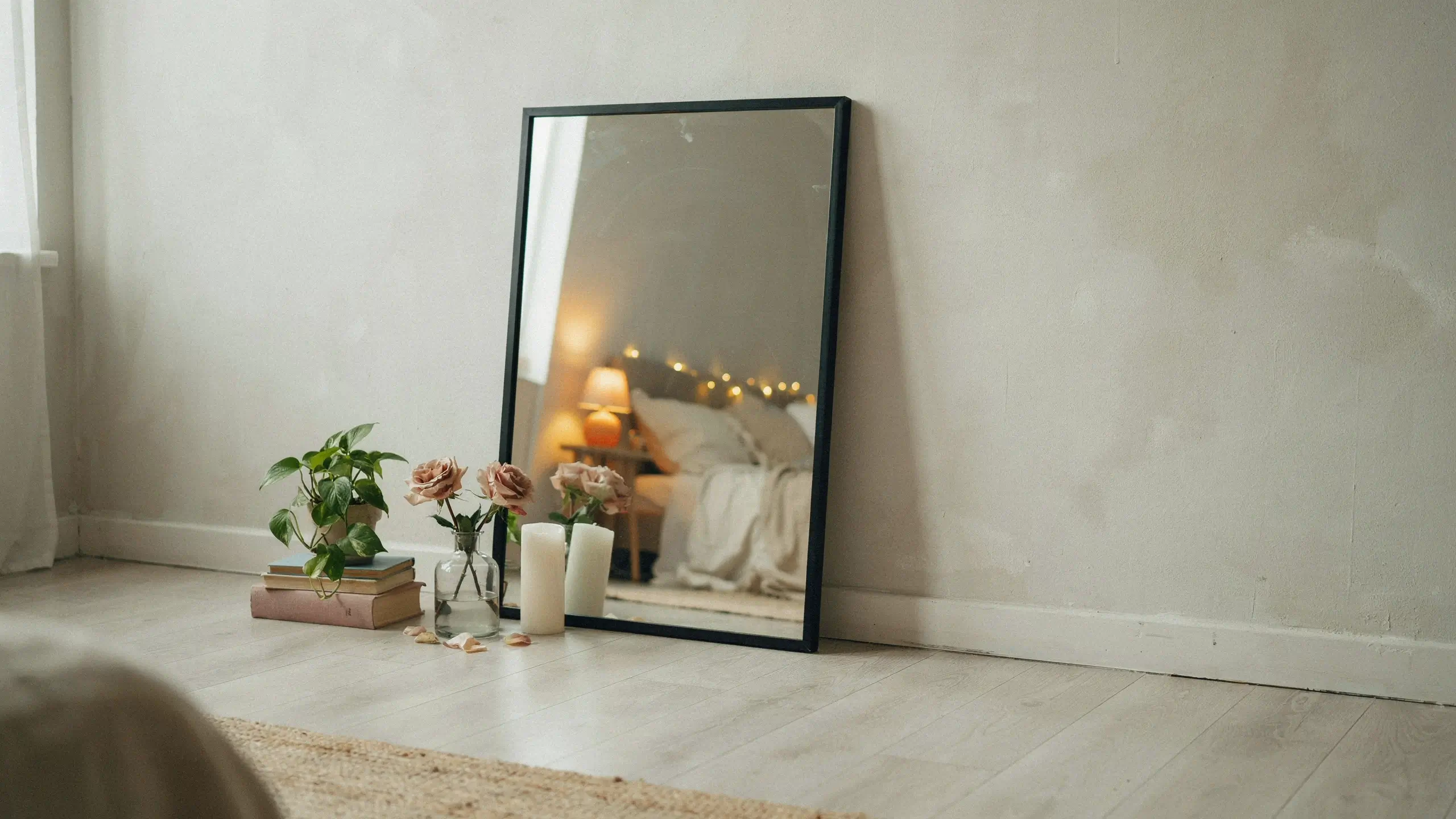 Black framed full-length mirror leaning on a white wall in a cozy bedroom, styled at the base with books, a green plant, pink roses in a vase, and white pillar candles.