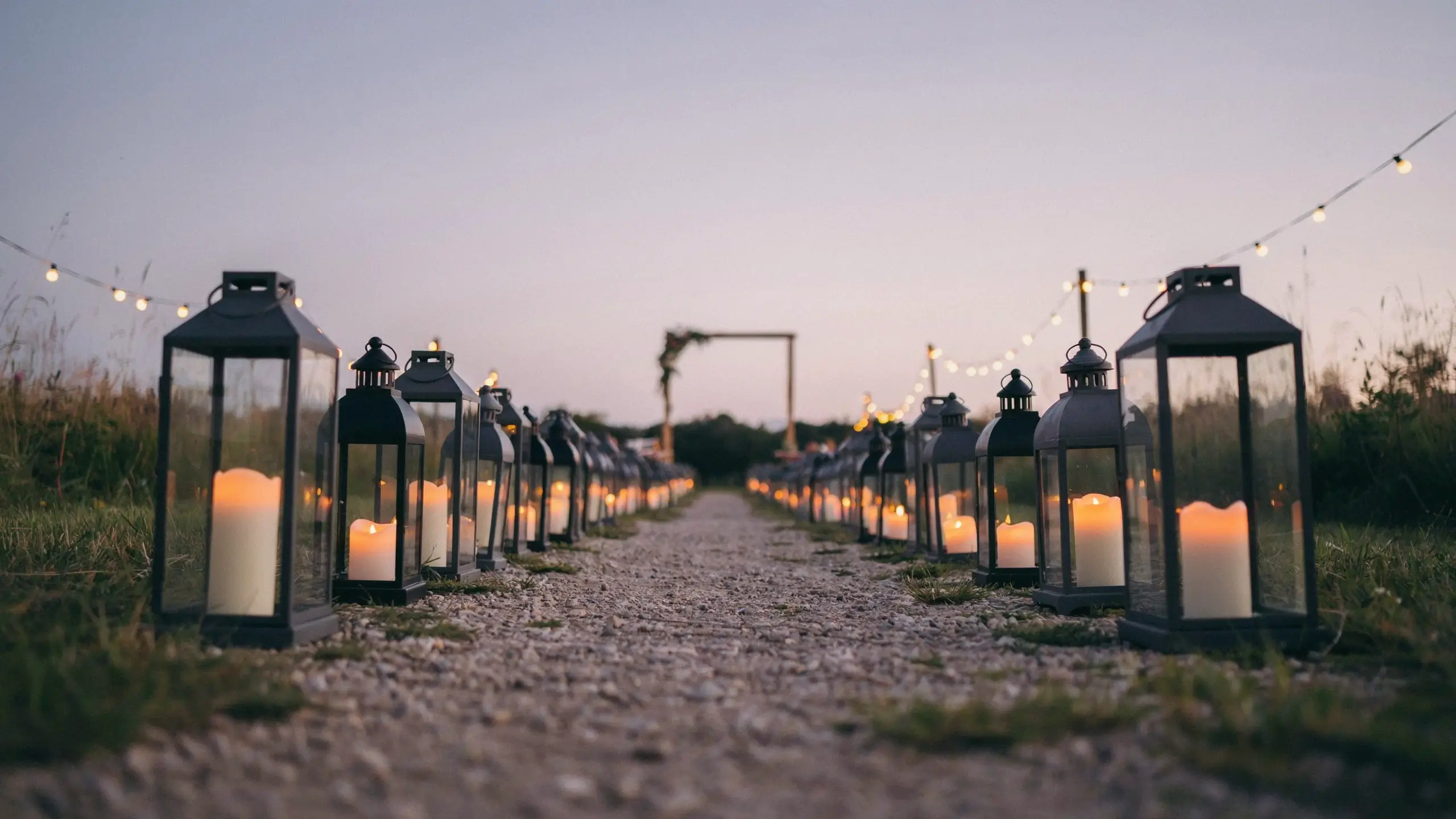 Black metal lanterns with white pillar candles lining a gravel wedding aisle during twilight with string lights overhead.