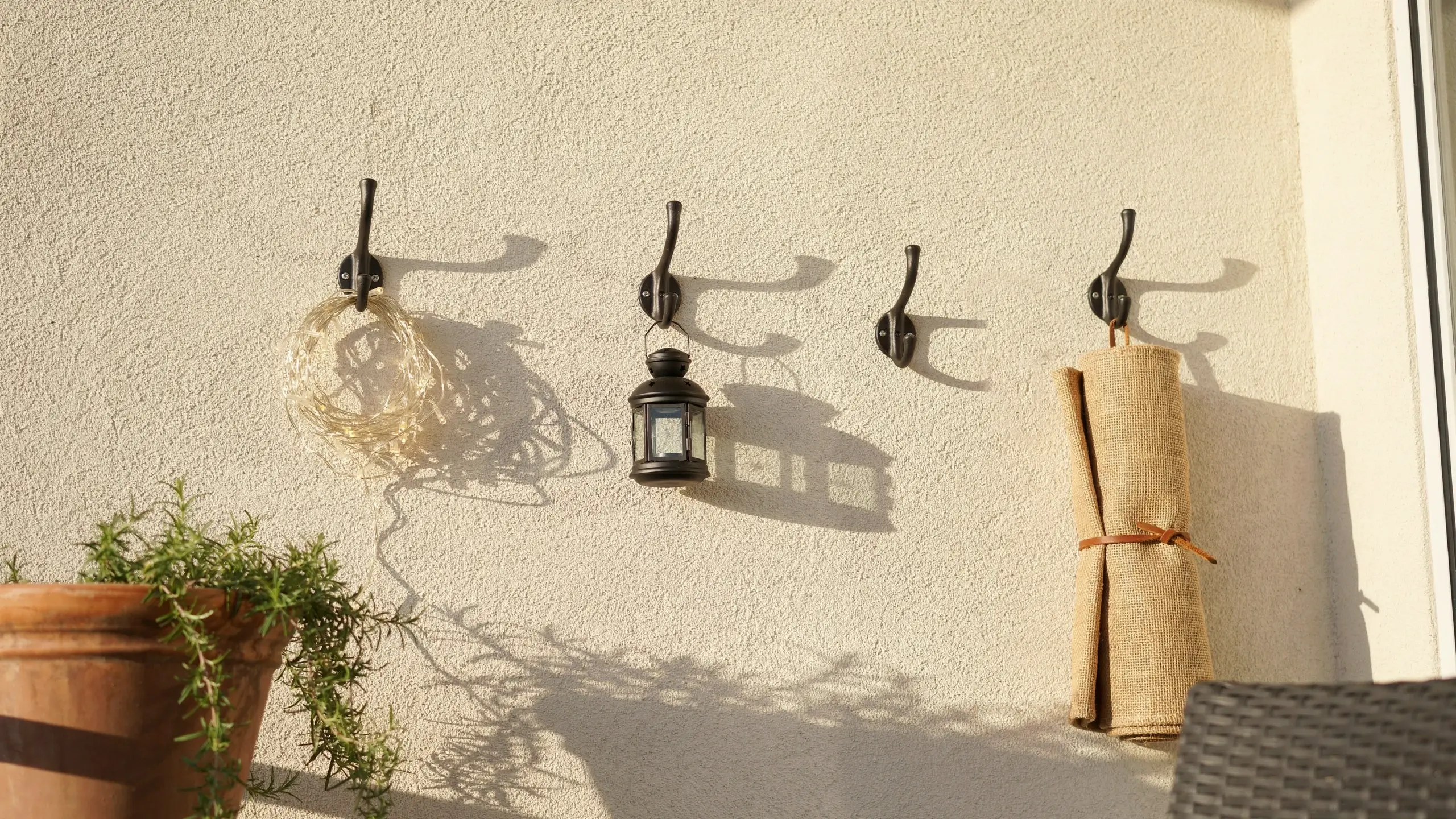 Four black metal outdoor wall hooks mounted on a white textured stucco wall, holding a coiled wire fairy light ring, a hanging black lantern, and a rolled burlap fabric tied with leather cord, with a terracotta potted herb plant visible on a sunny patio.