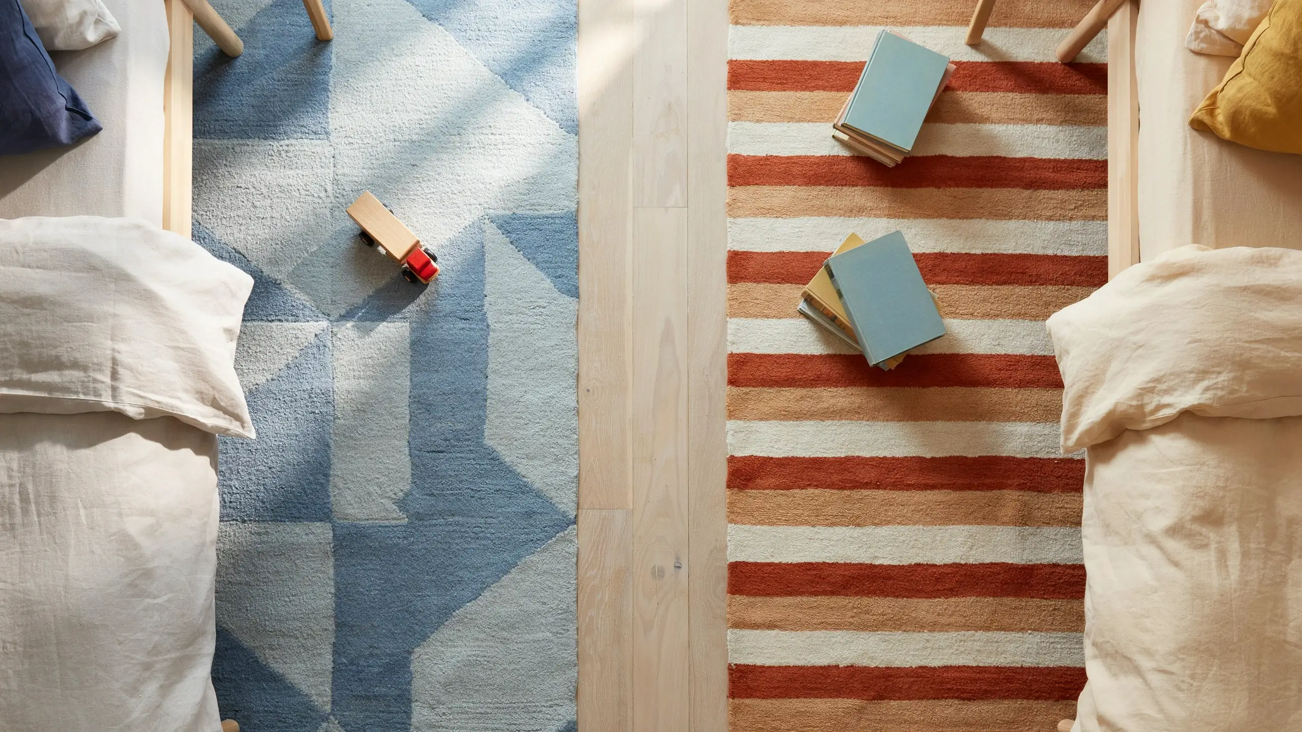 Top-down view of a shared children's bedroom with two rugs instead of one—a blue geometric rug and a striped orange rug—separating two twin beds with pillows, books, and a toy truck.
