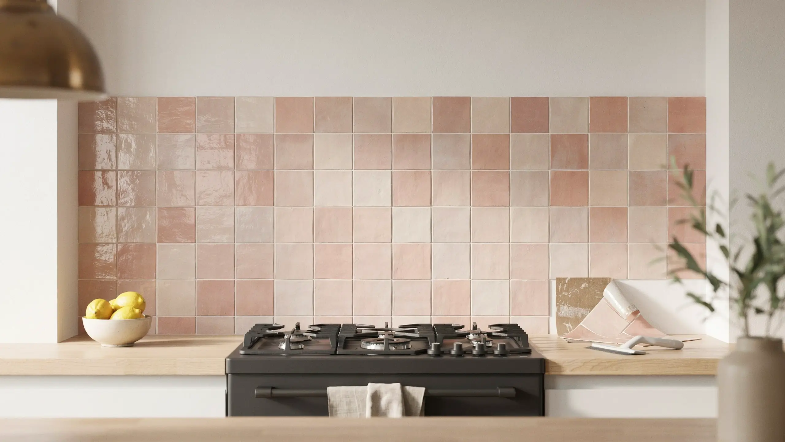 Blush pink square peel-and-stick tile backsplash installed above a matte black gas stove, featuring a bowl of fresh lemons and a brass pendant light in a modern kitchen renovation.