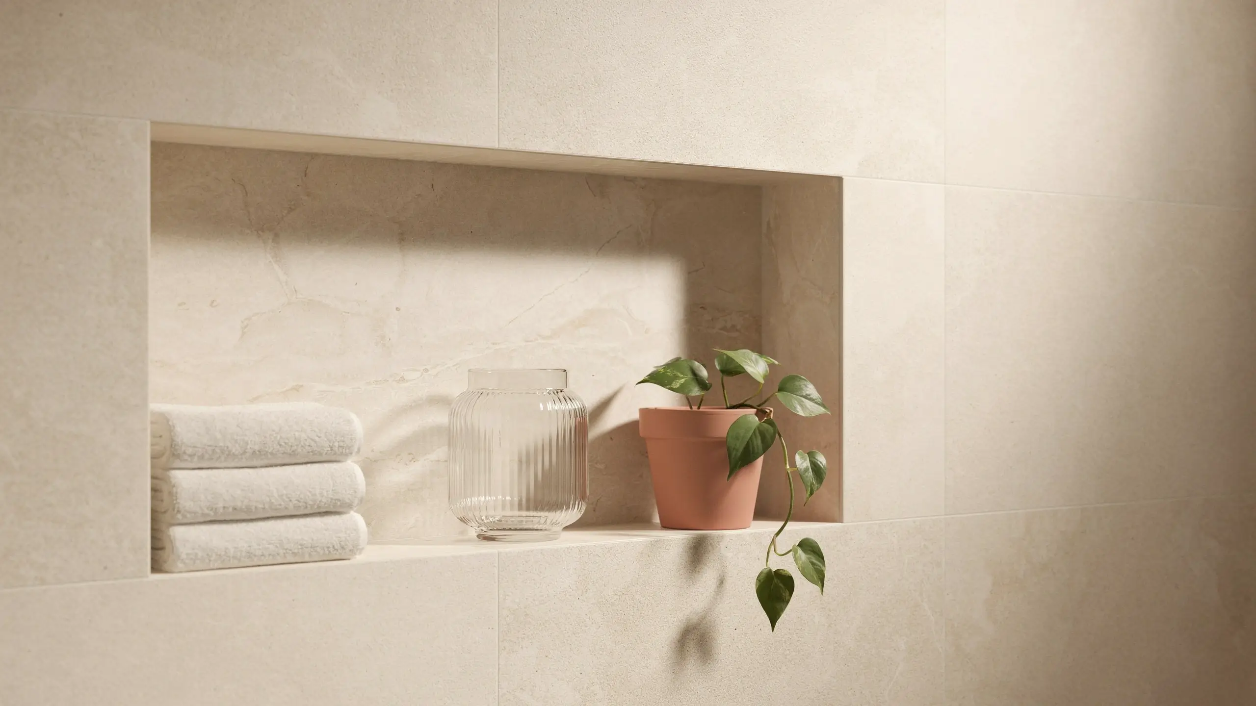 Minimalist beige marble bathroom niche styled with folded white towels, a ribbed glass jar, and a trailing green pothos plant in a terracotta pot, creating a boutique hotel spa aesthetic.