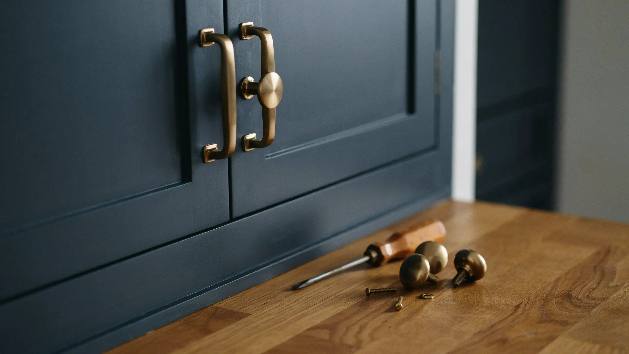 Dark blue kitchen cabinets featuring new brushed brass curved pull handles and a round knob, with a wooden-handled screwdriver and spare knobs resting on a warm oak countertop during a DIY hardware swap.