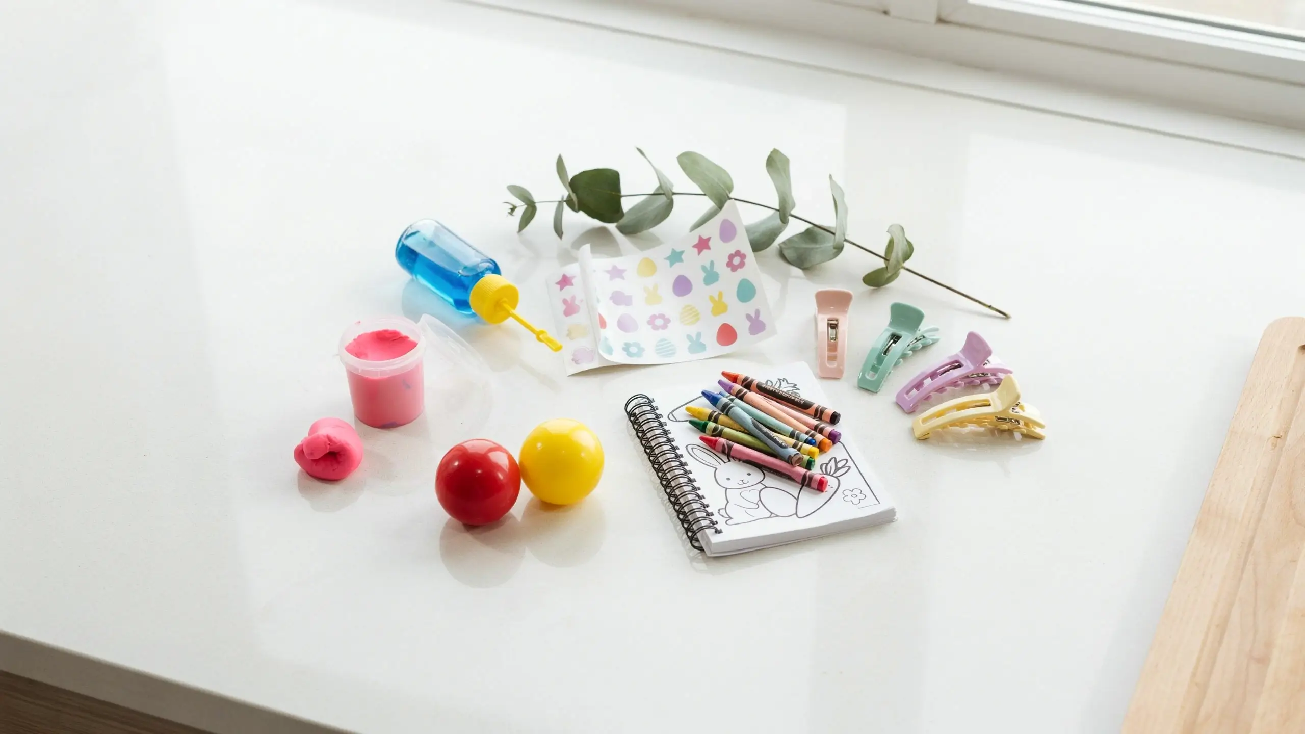 Flat lay of dollar store Easter basket filler ideas including a bunny coloring notebook, crayons, spring-themed stickers, pink playdough, pastel hair clips, and colorful plastic eggs on a white countertop.