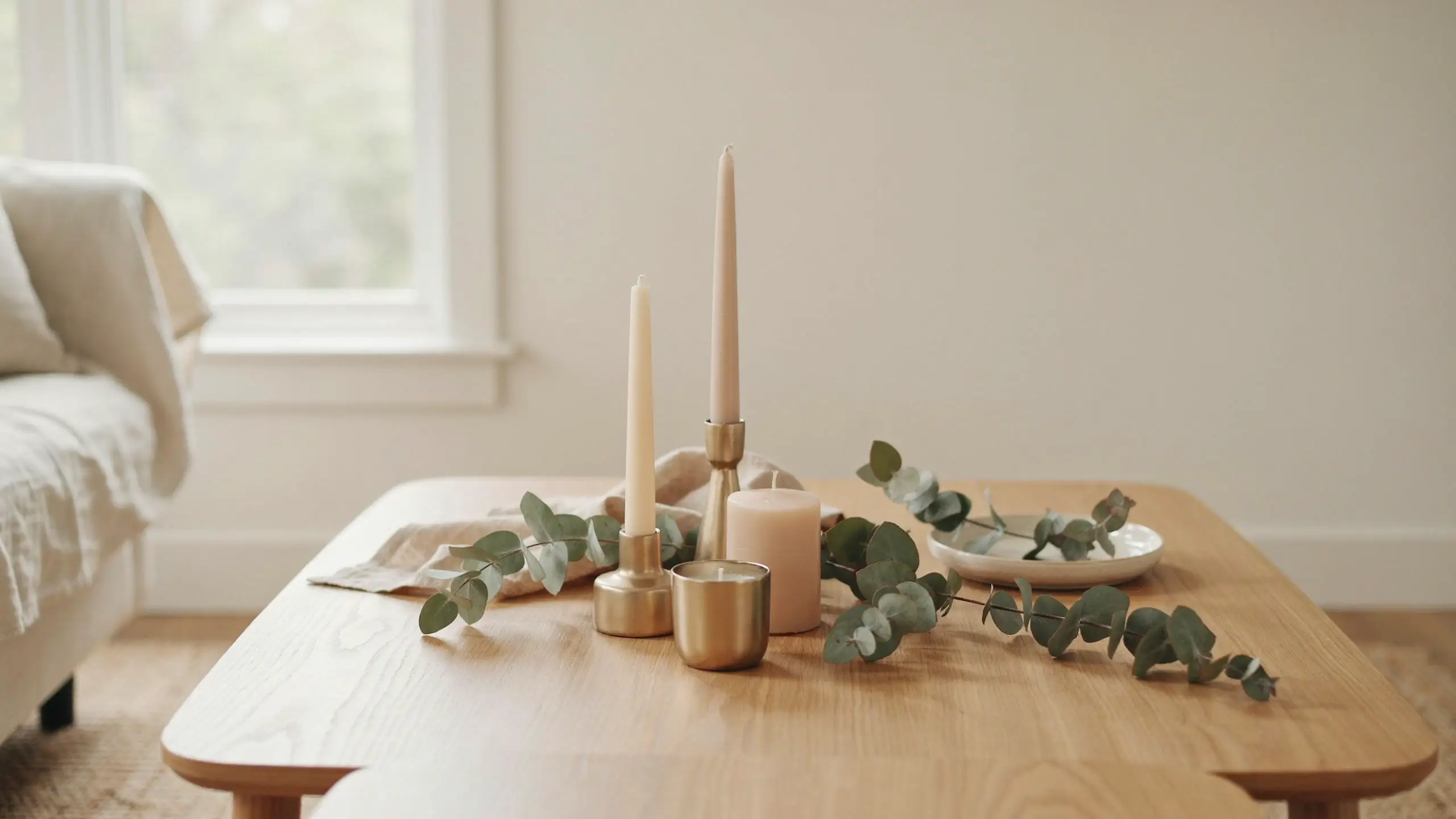 Minimalist coffee table arrangement featuring champagne gold candle holders with cream taper candles, a blush pillar candle, and fresh sage green eucalyptus sprigs on a light oak wood table in a bright, cozy living room.