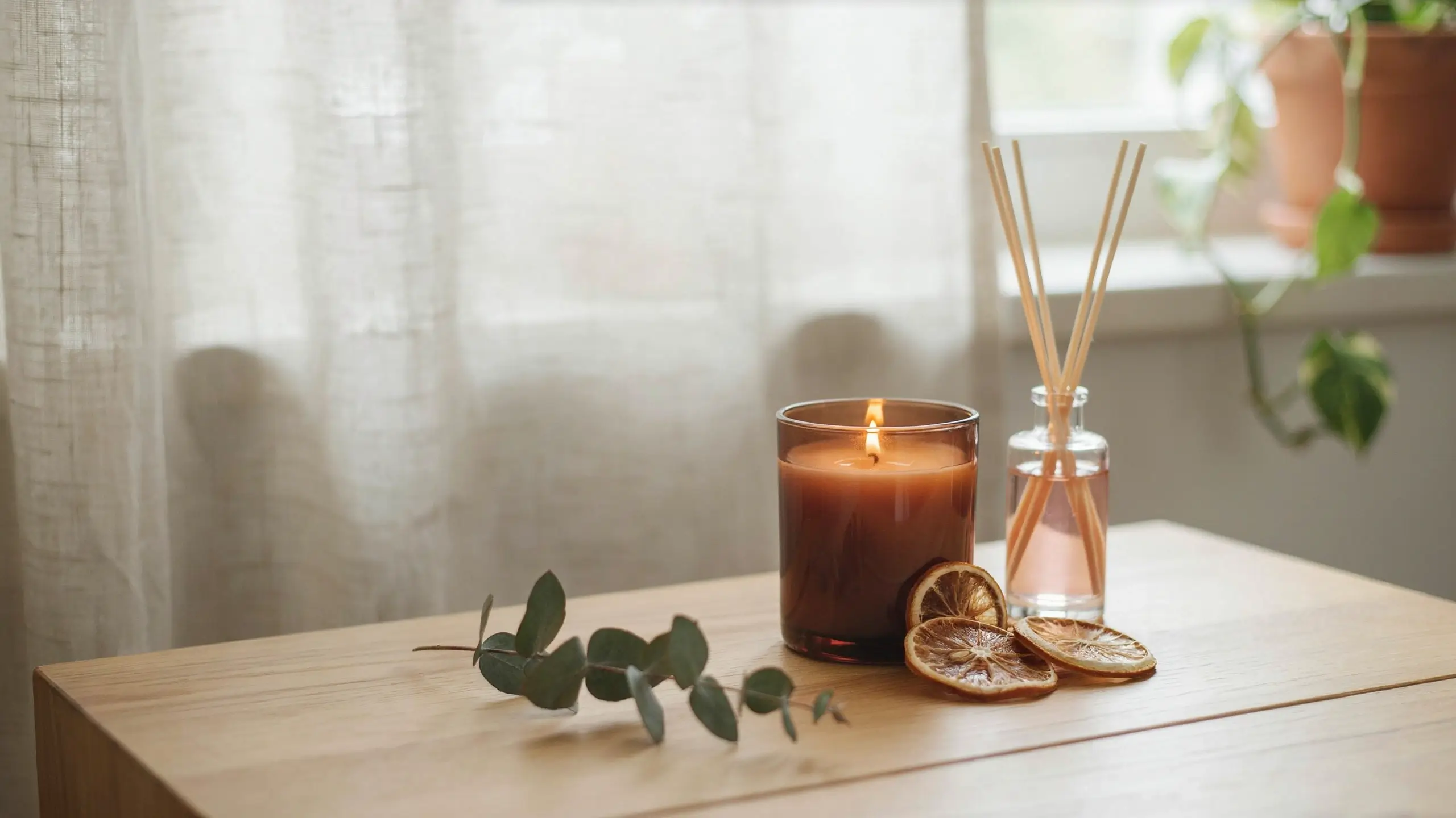 A cozy home scent change setup with a lit amber candle, glass reed diffuser, dried orange slices, and eucalyptus leaves on a light wooden table.