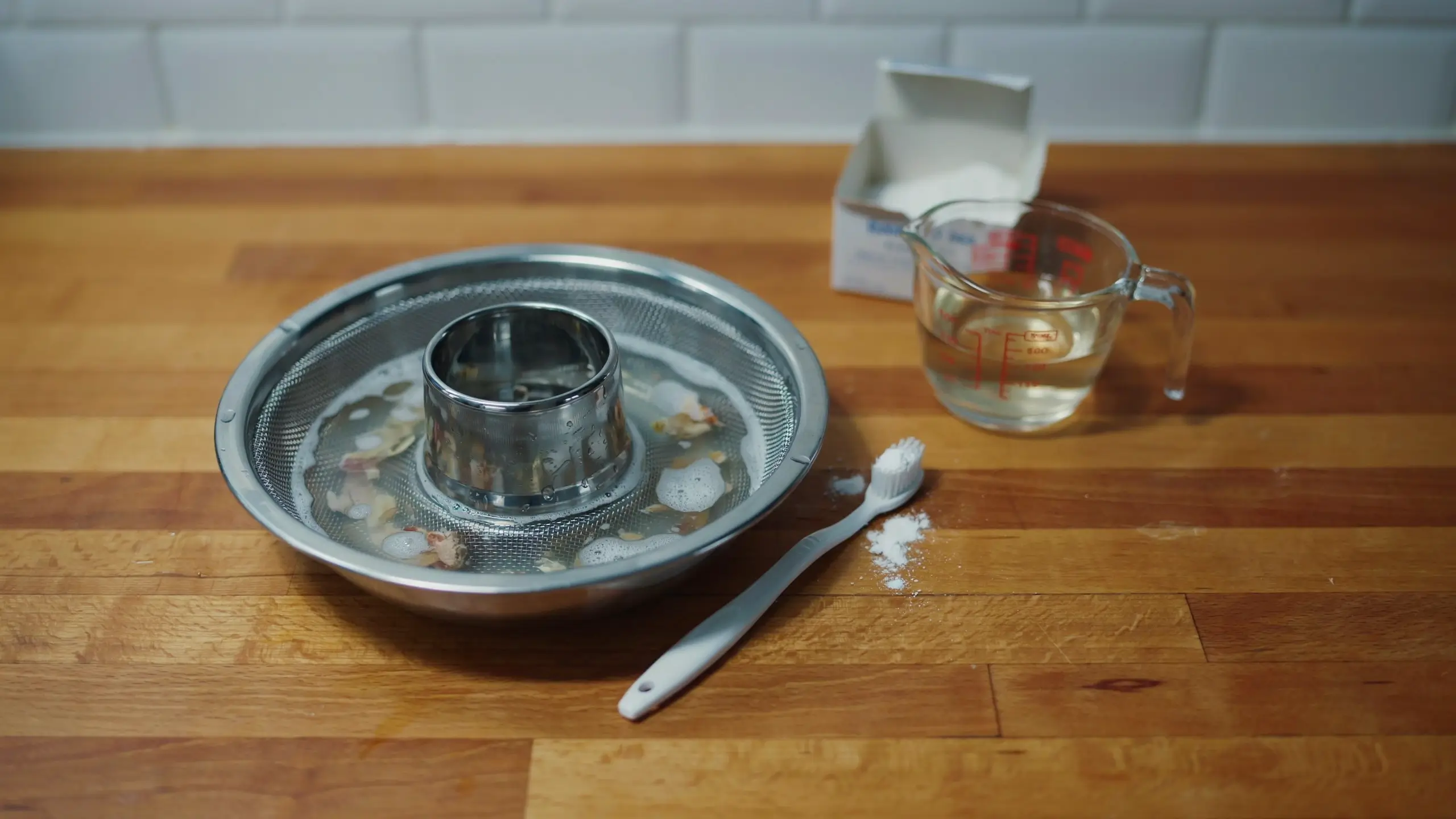Close-up of a stainless steel mesh filter soaking in a shallow bowl of soapy water, surrounded by a white cleaning toothbrush and baking soda on a wooden kitchen countertop.