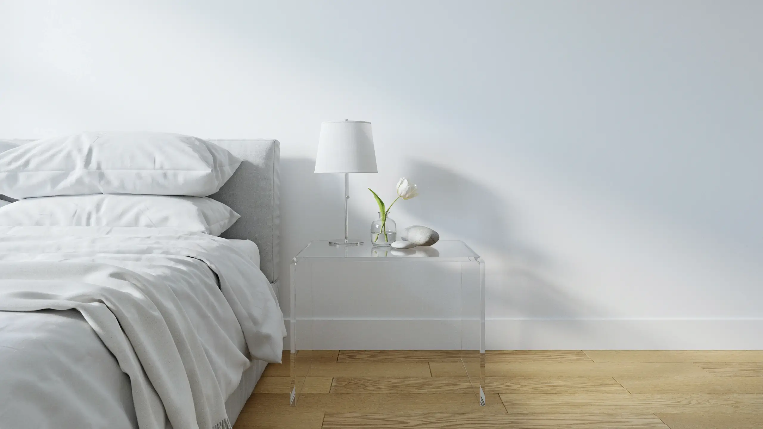 Slim acrylic ghost nightstand in a minimalist white bedroom, styling a modern lamp and a fresh white tulip. 