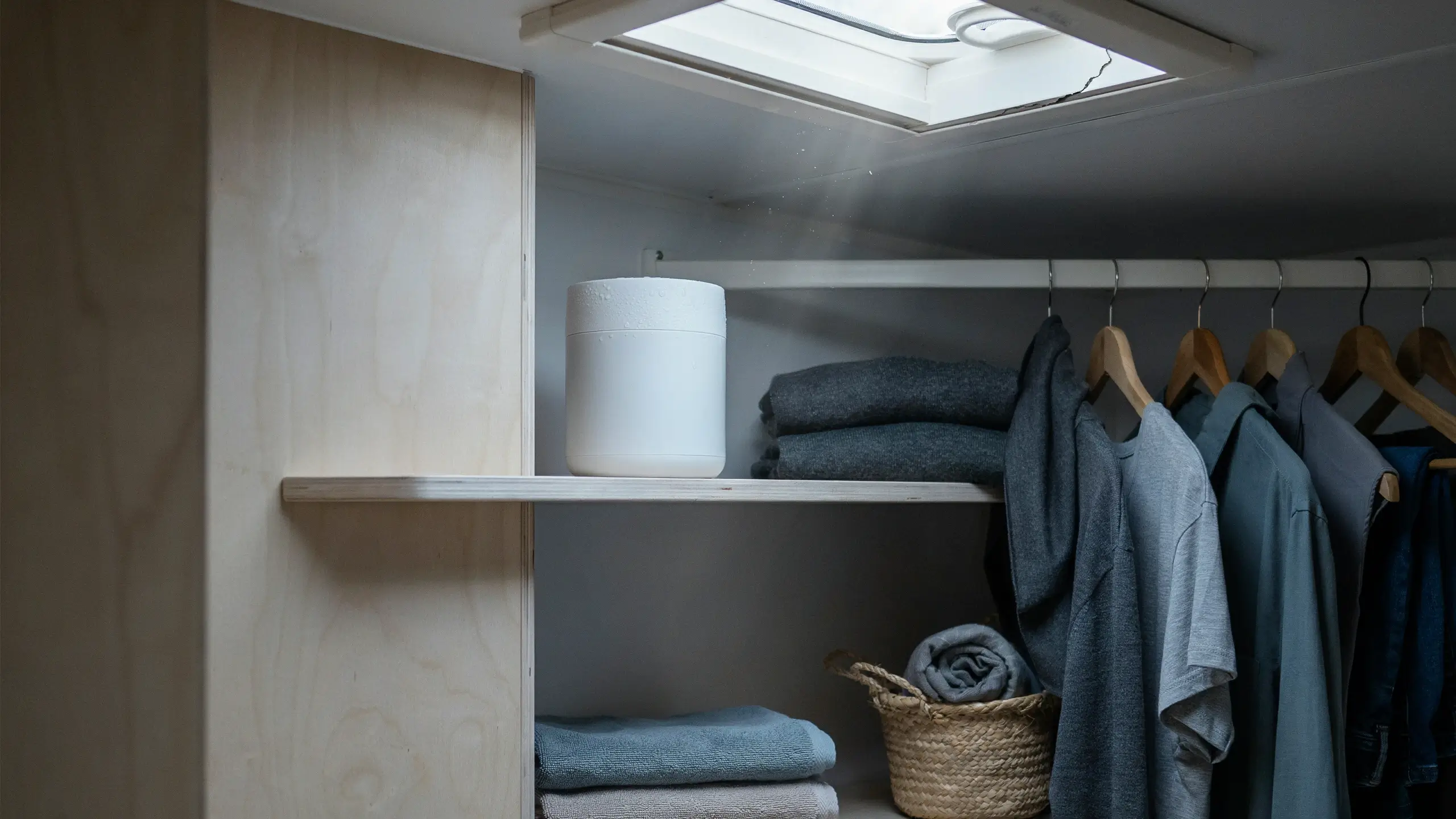 White dehumidifier on a wooden shelf inside a closet controlling moisture to protect hanging clothes and folded towels.